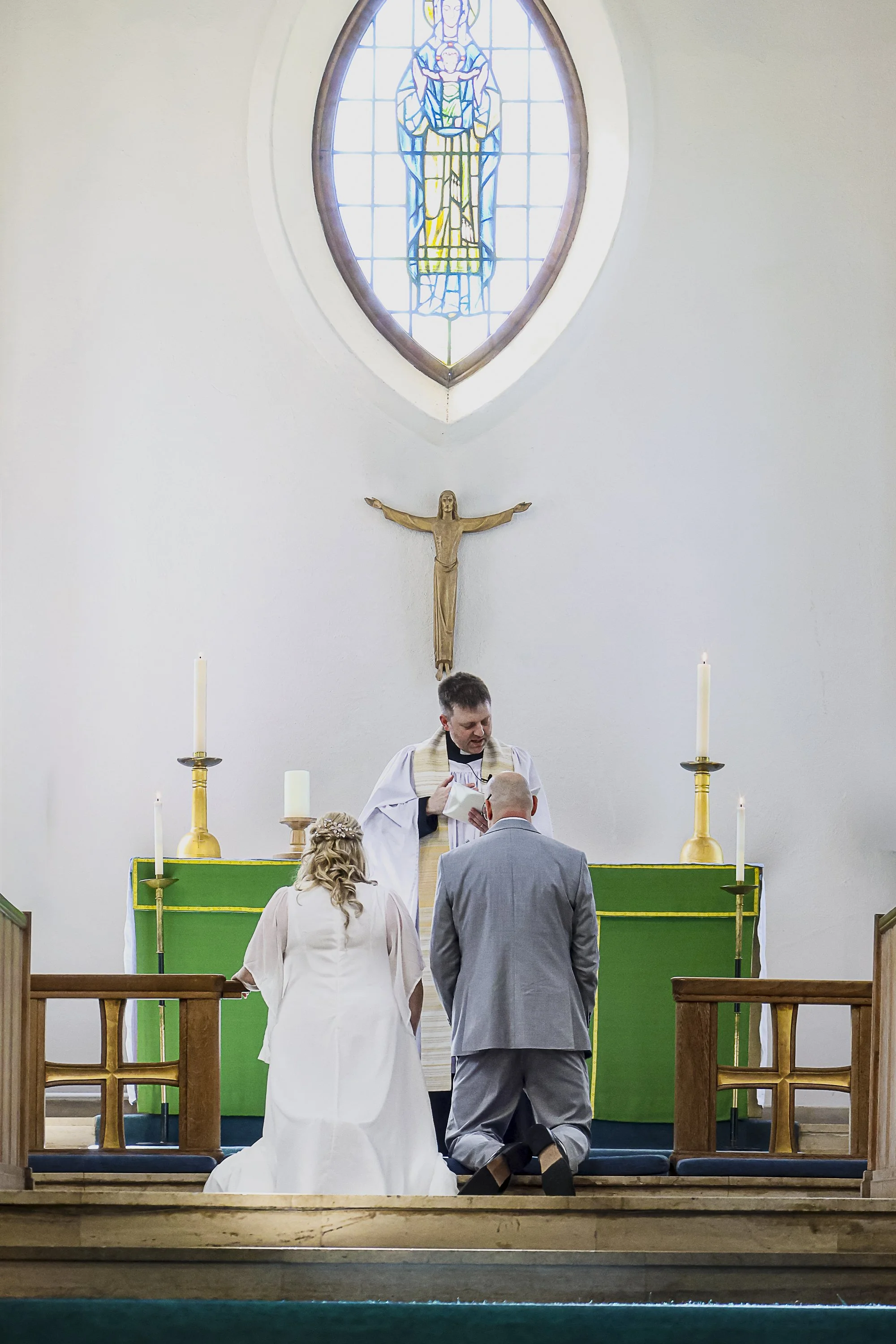 A wedding ceremony in a church with the bride and groom kneeling before the priest. The background includes a stained glass window of Mary holding baby Jesus, and a crucifix on the wall. Candles are on the altar.