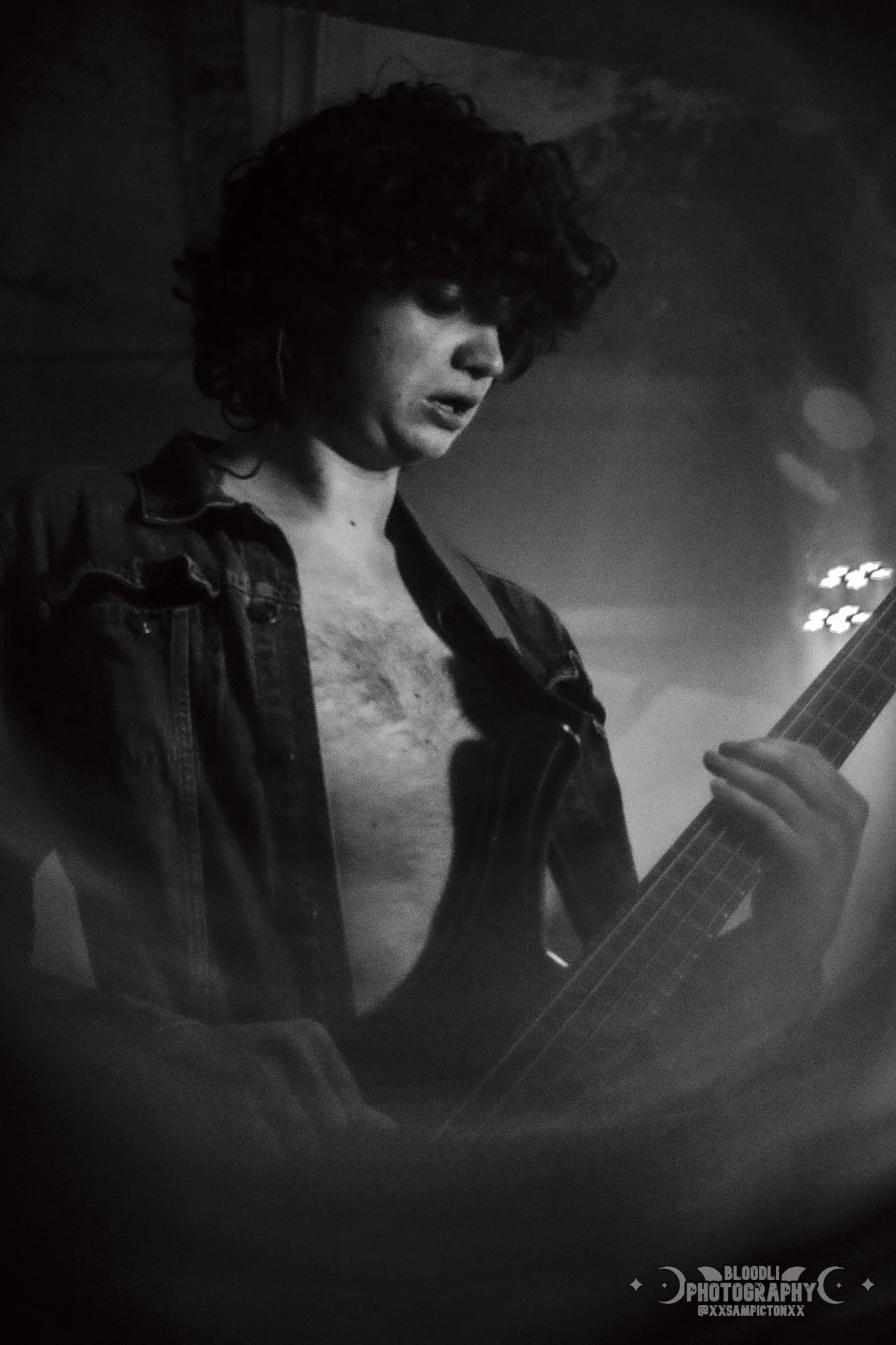 Black and white photo of a woman with curly hair wearing an open denim jacket, playing an electric guitar.