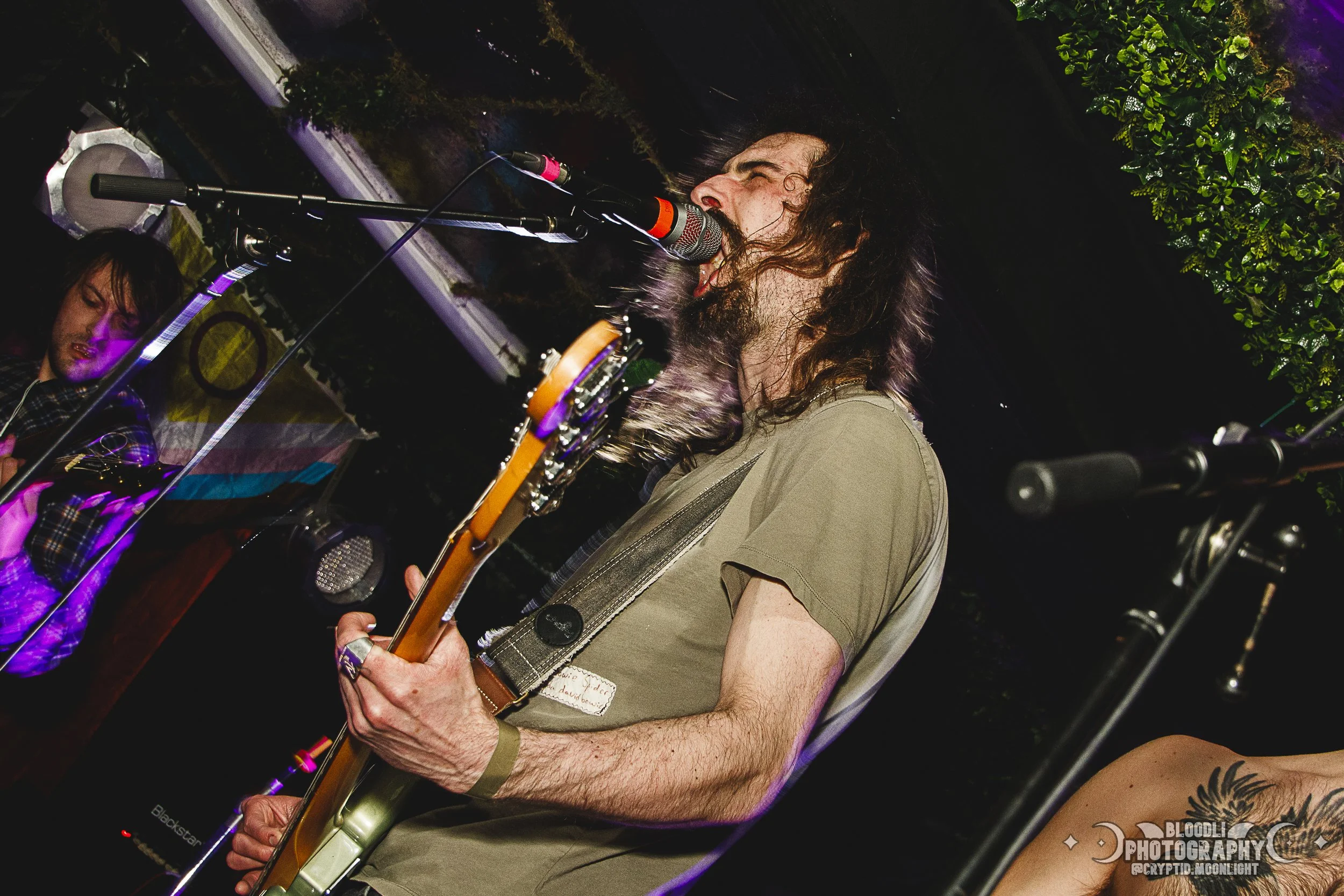Musician singing into microphone while playing electric guitar at a live outdoor concert.