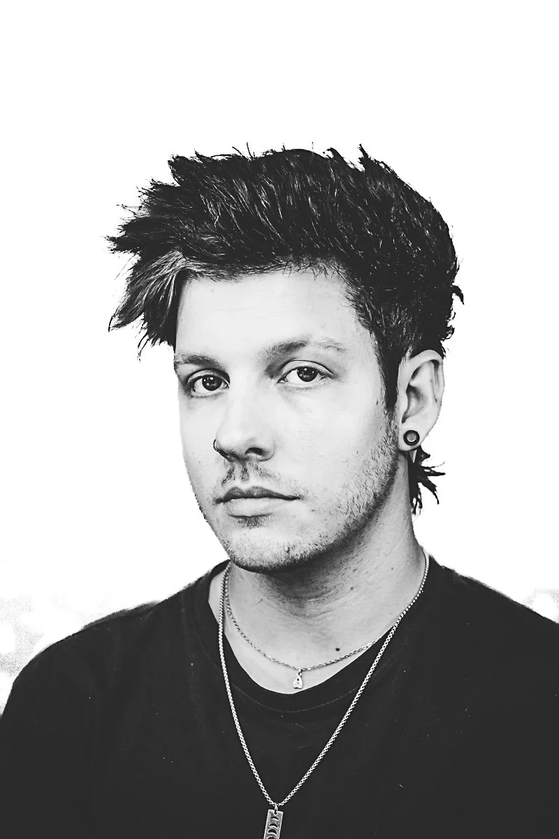Black and white portrait of a young man with short, tousled hair, earrings, and layered necklaces, wearing a dark shirt.