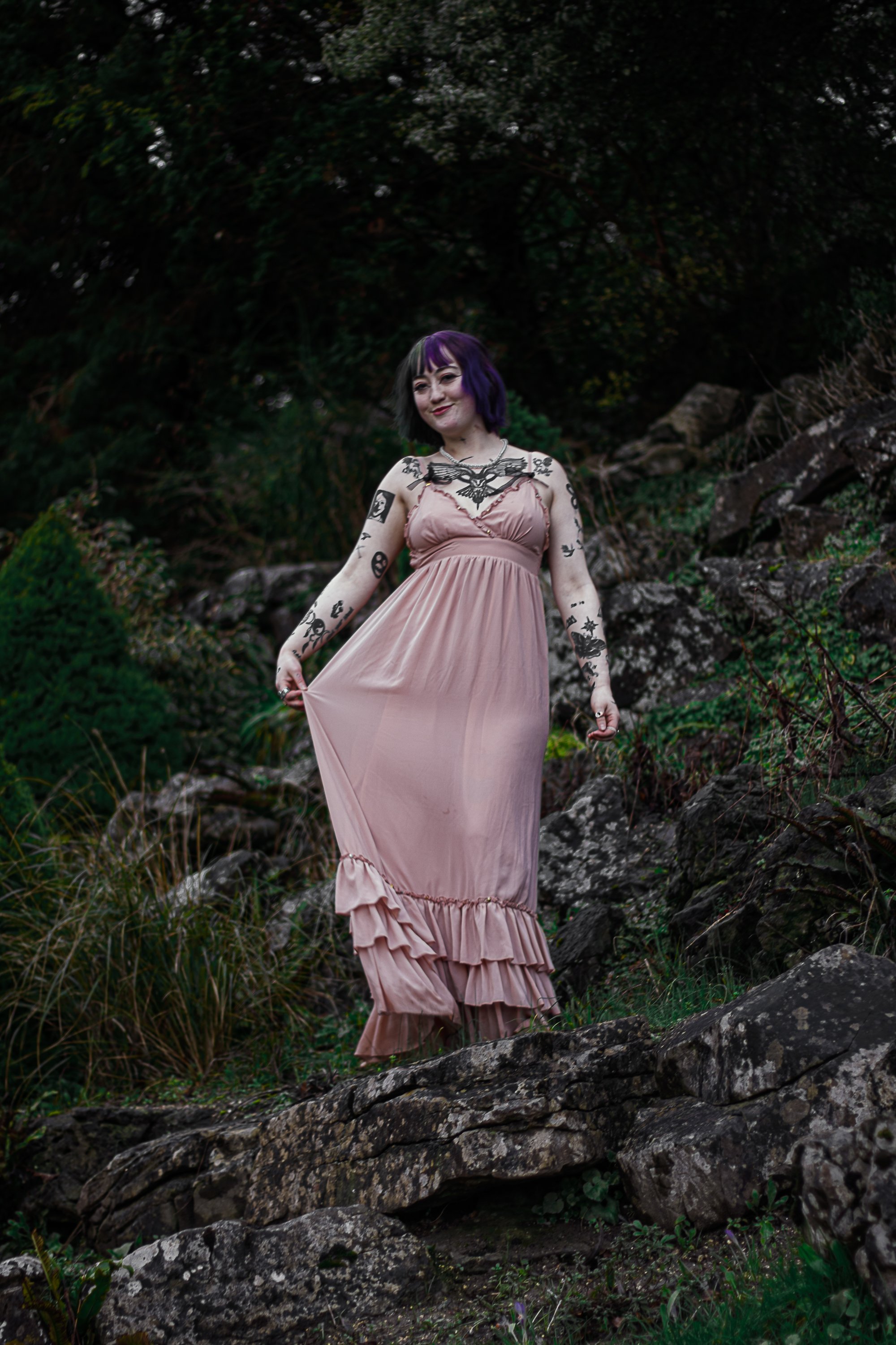A young woman with purple and black hair wearing a pink dress with ruffles is walking outdoors on rocks and grass, surrounded by greenery.