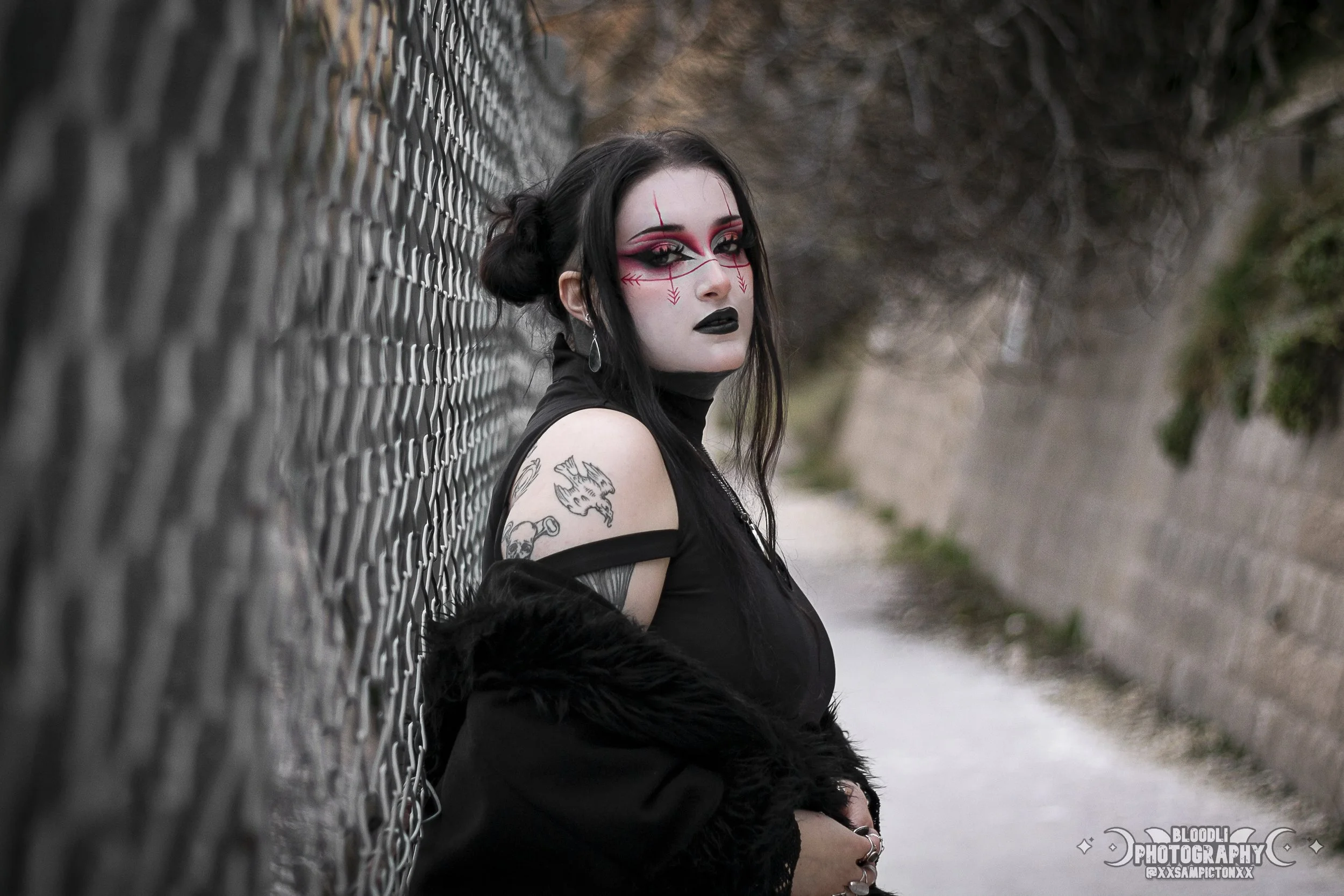 A woman with gothic makeup and tattoos leaning against a chain-link fence outdoors.