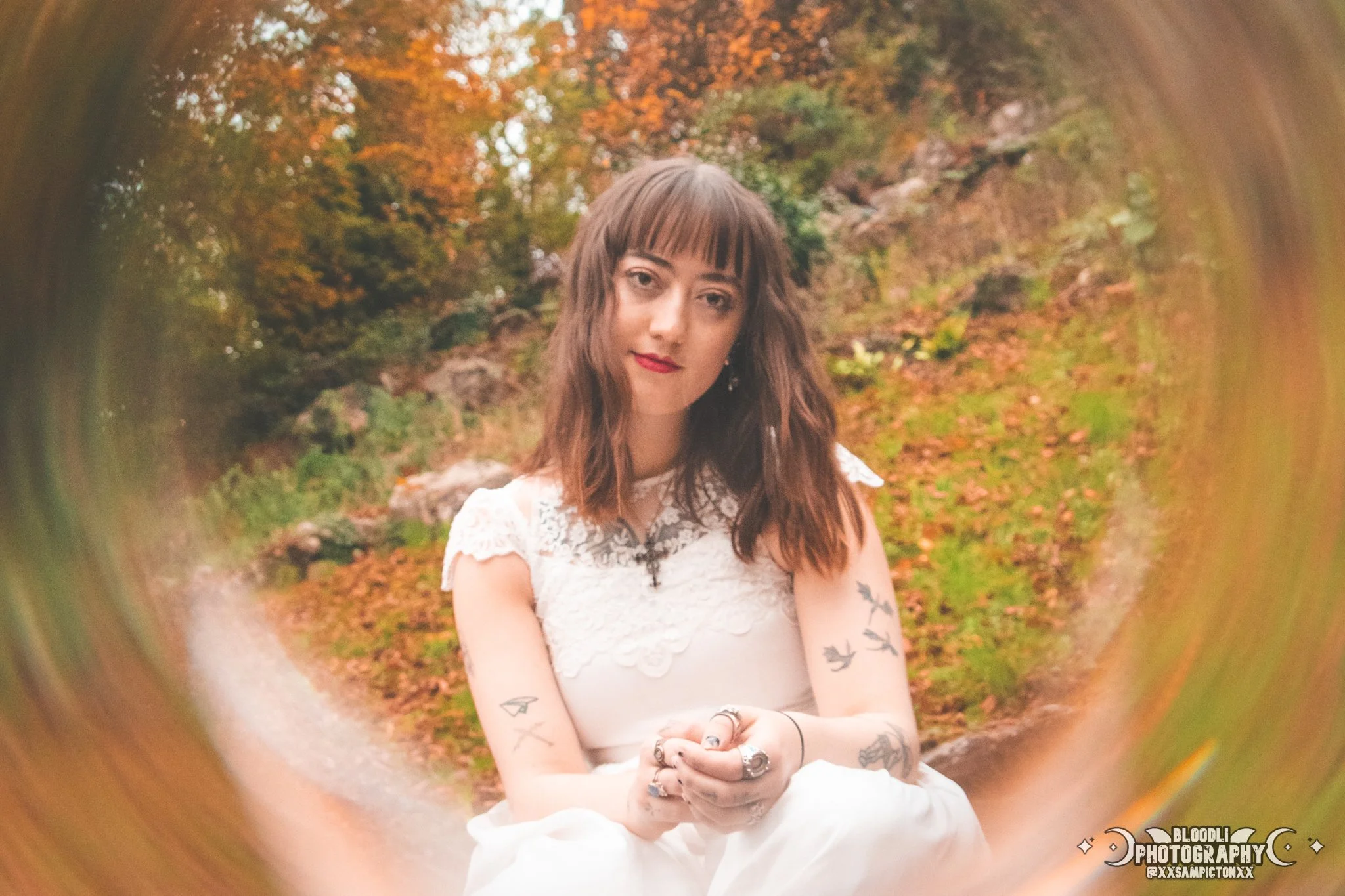A young woman with long brown hair and tattoos on her arms sits outdoors in a forest with autumn leaves. She wears a white lace dress and has red lipstick. The photo is taken through a circular frame, creating a tunnel effect.