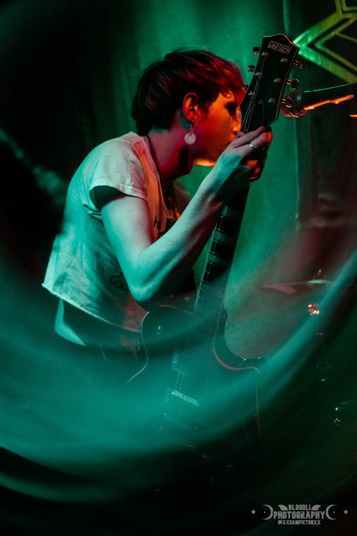 A woman with short dark hair, wearing earrings and a light-colored top, is performing on stage with a guitar, surrounded by colorful stage lighting.