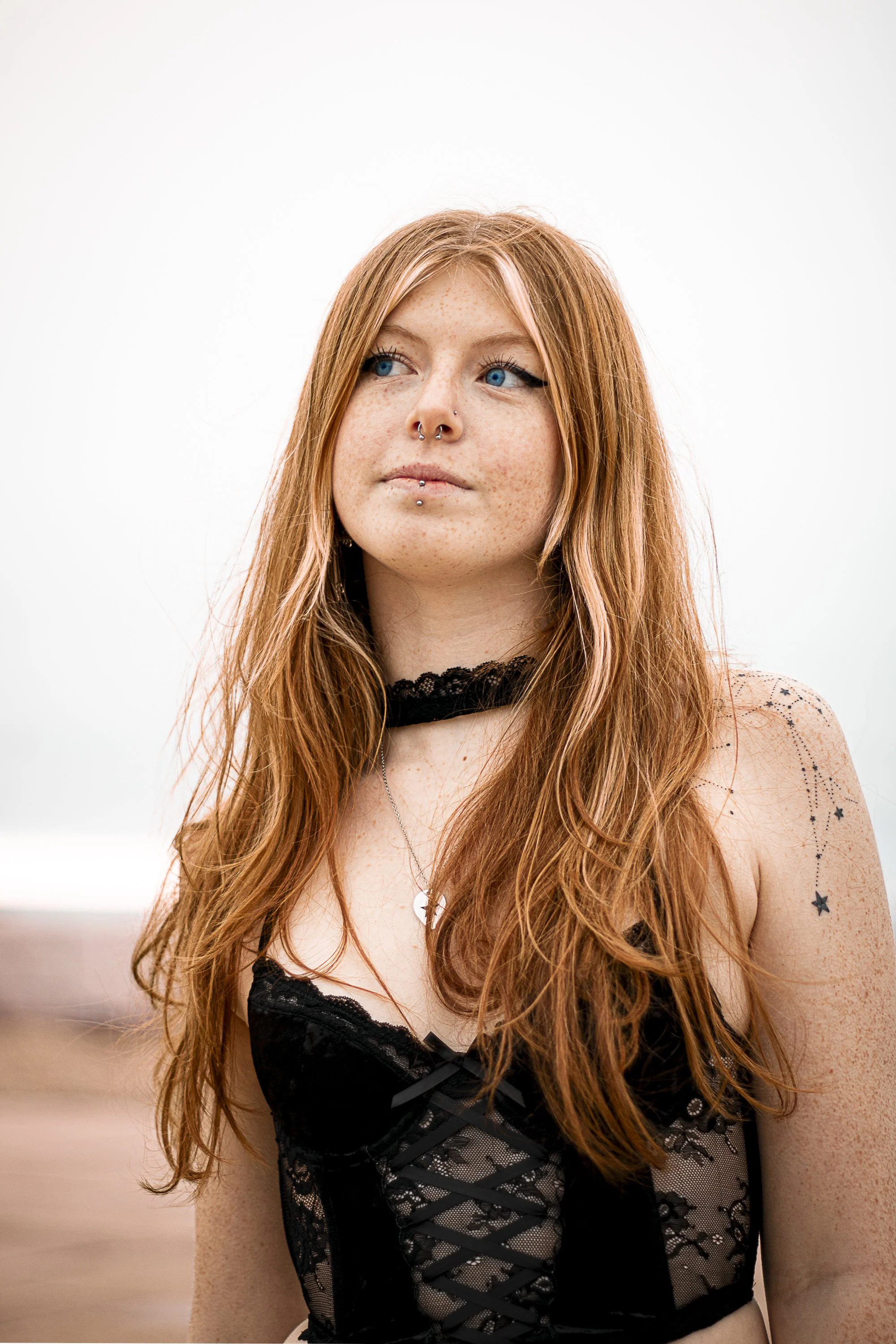 A young woman with long red hair, blue eyes, and multiple facial piercings, wearing a black lace top and a choker, standing against a plain background.