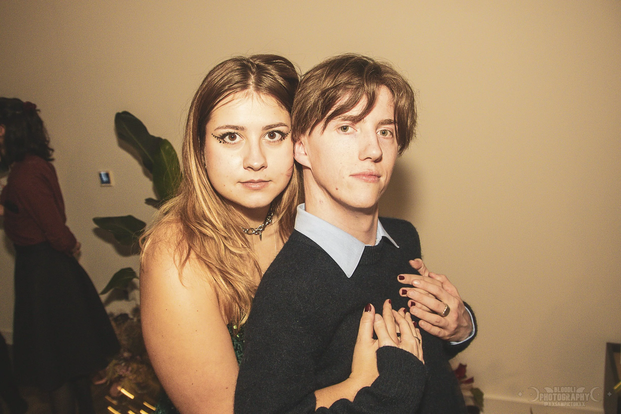 A young woman with long, wavy hair and makeup, and a young man with short, tousled hair pose closely together, with the woman's arms wrapped around the man's shoulders and her hands on his chest. They are looking directly at the camera, in an indoor 
