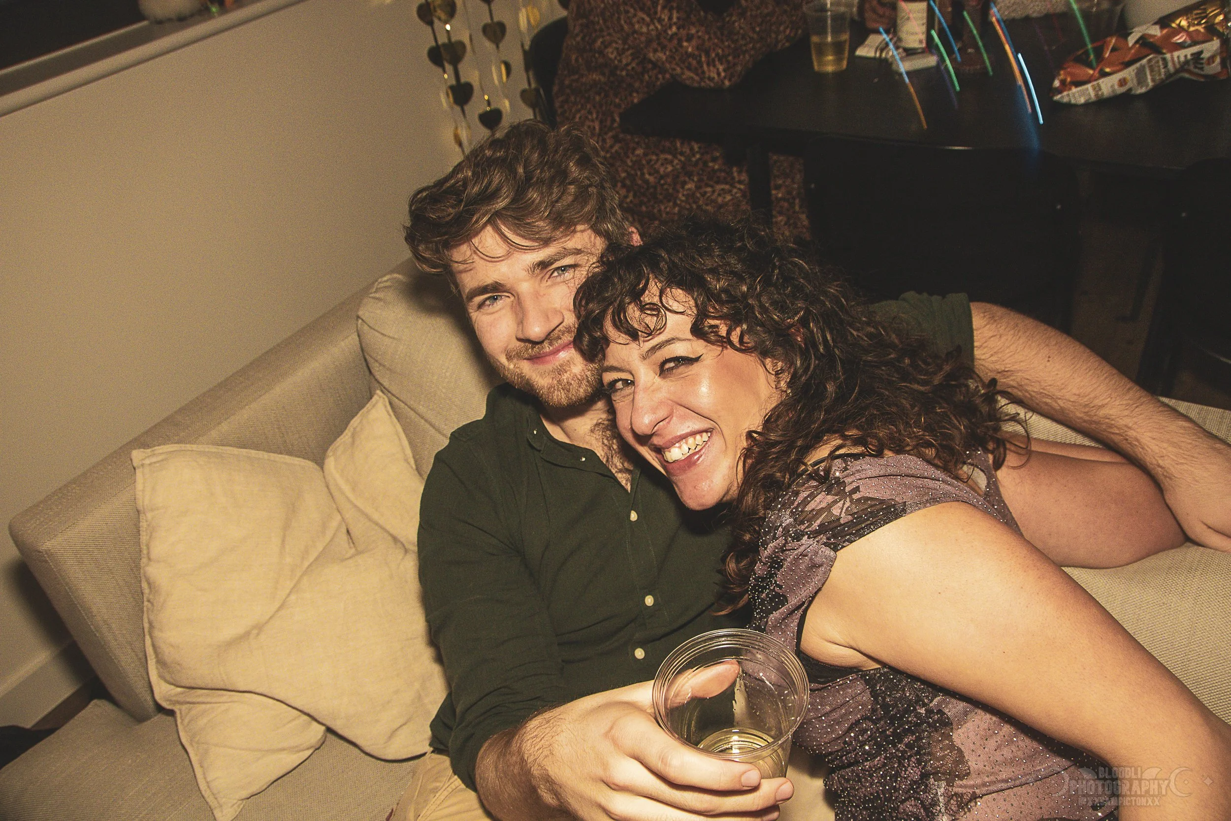 A man and a woman sitting on a beige sofa, holding a drink, smiling at the camera at a social gathering or party.