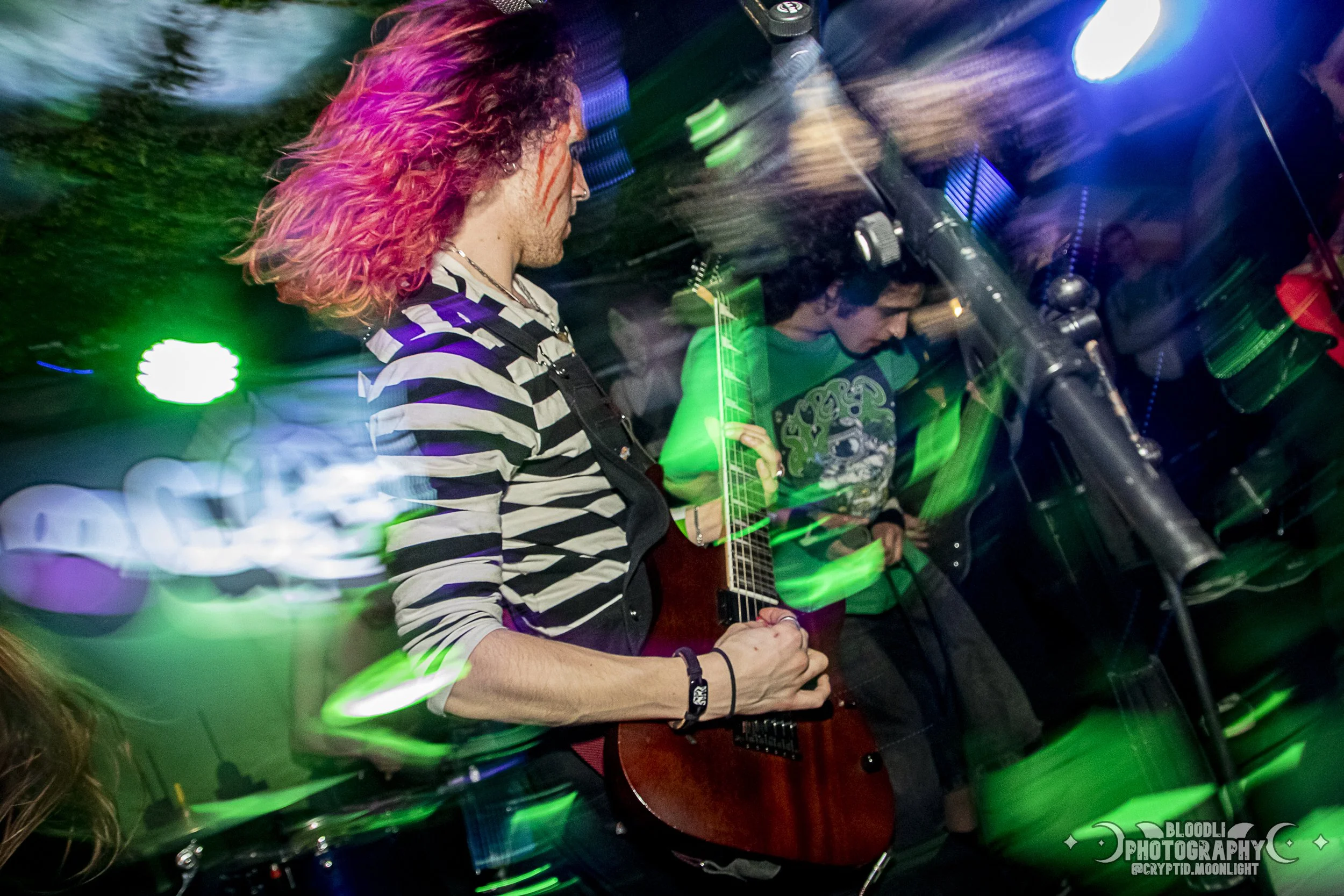 A person with curly hair dyed pink and purple wearing a black and white striped shirt playing an electric guitar at a concert with colorful lights and motion blur.