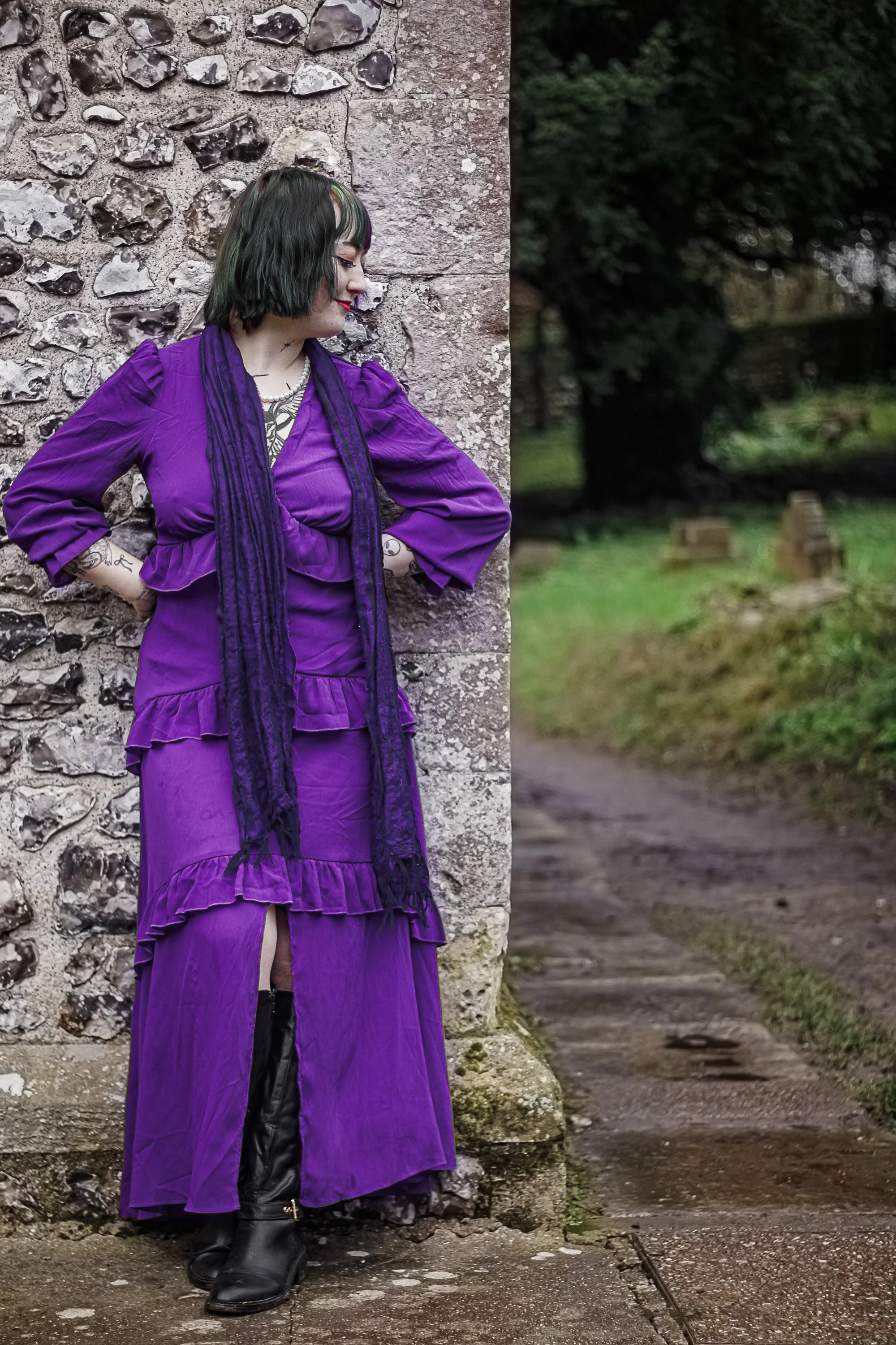 A woman with black hair with green streaks, dressed in a purple outfit, standing against a stone wall in an outdoor setting with trees and a path in the background.