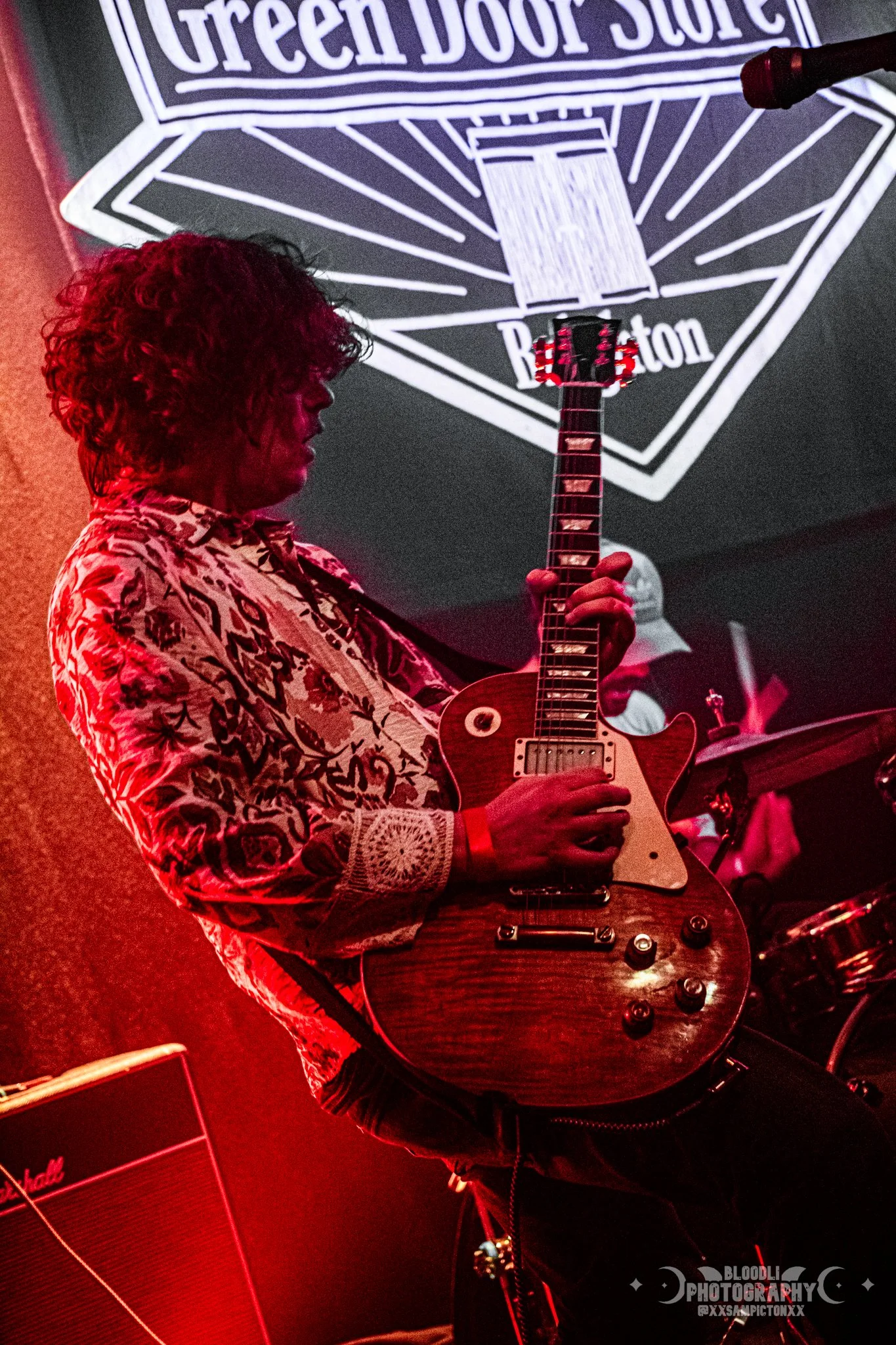 A musician playing an electric guitar on stage with a large sign in the background that reads 'Green Door Store' and 'Brighton'.