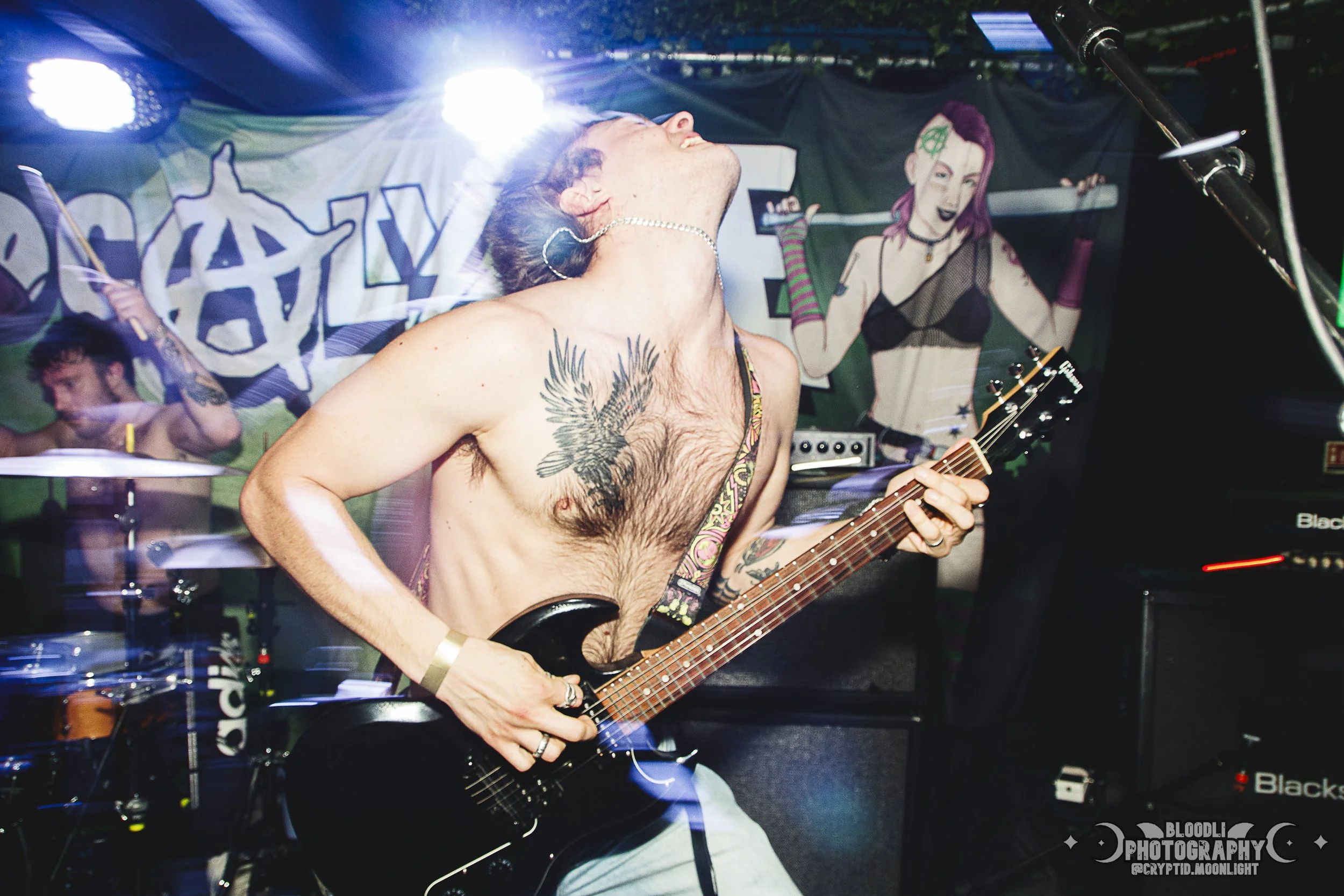 A shirtless guitarist with tattoos and earrings passionately performing on stage at a concert. There is a drum set behind him and a colorful banner in the background.