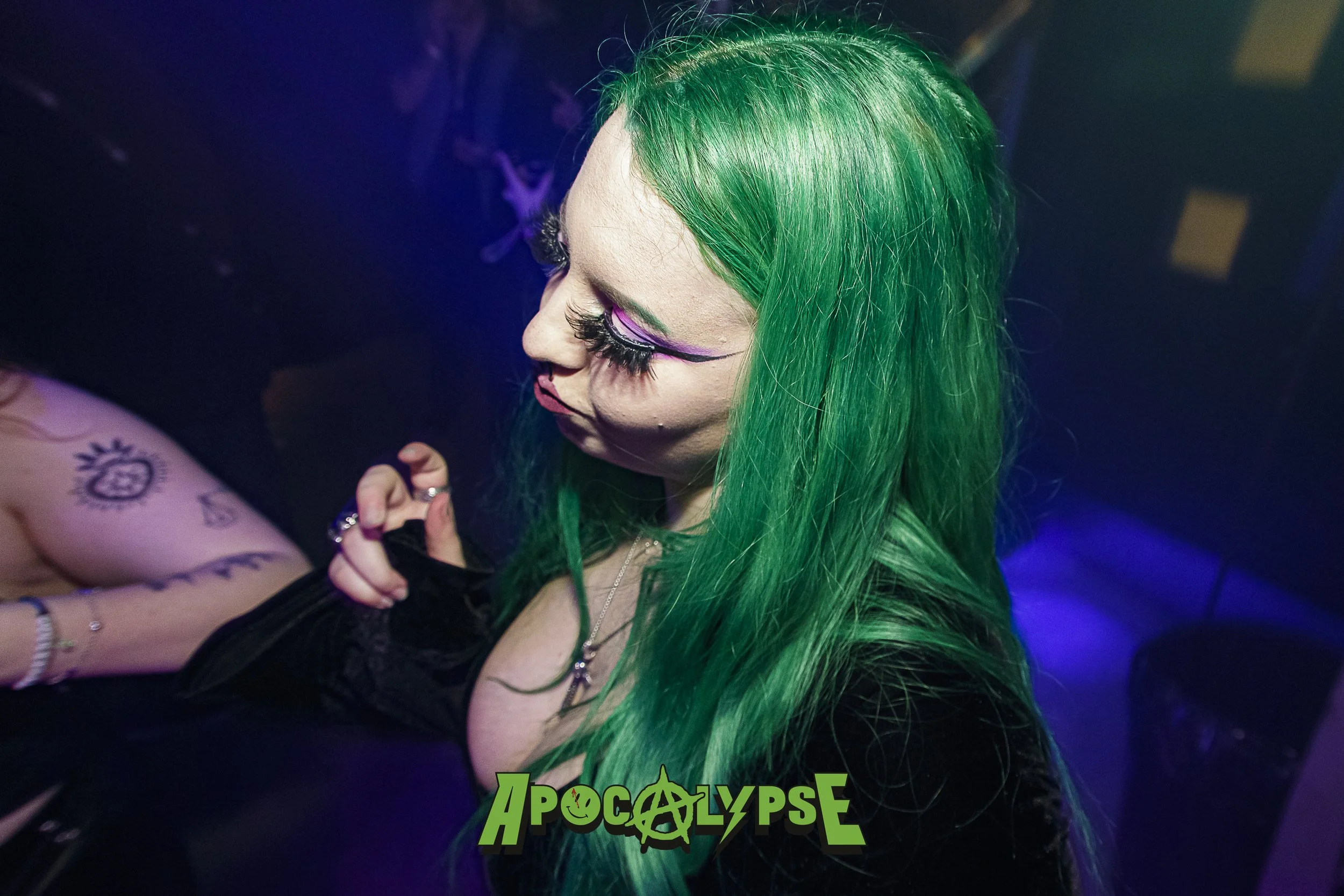 A woman with long, bright green hair and dramatic makeup, including bold eyelashes and purple eyeshadow, in a dark setting with blue and purple lighting, at an event or nightclub.