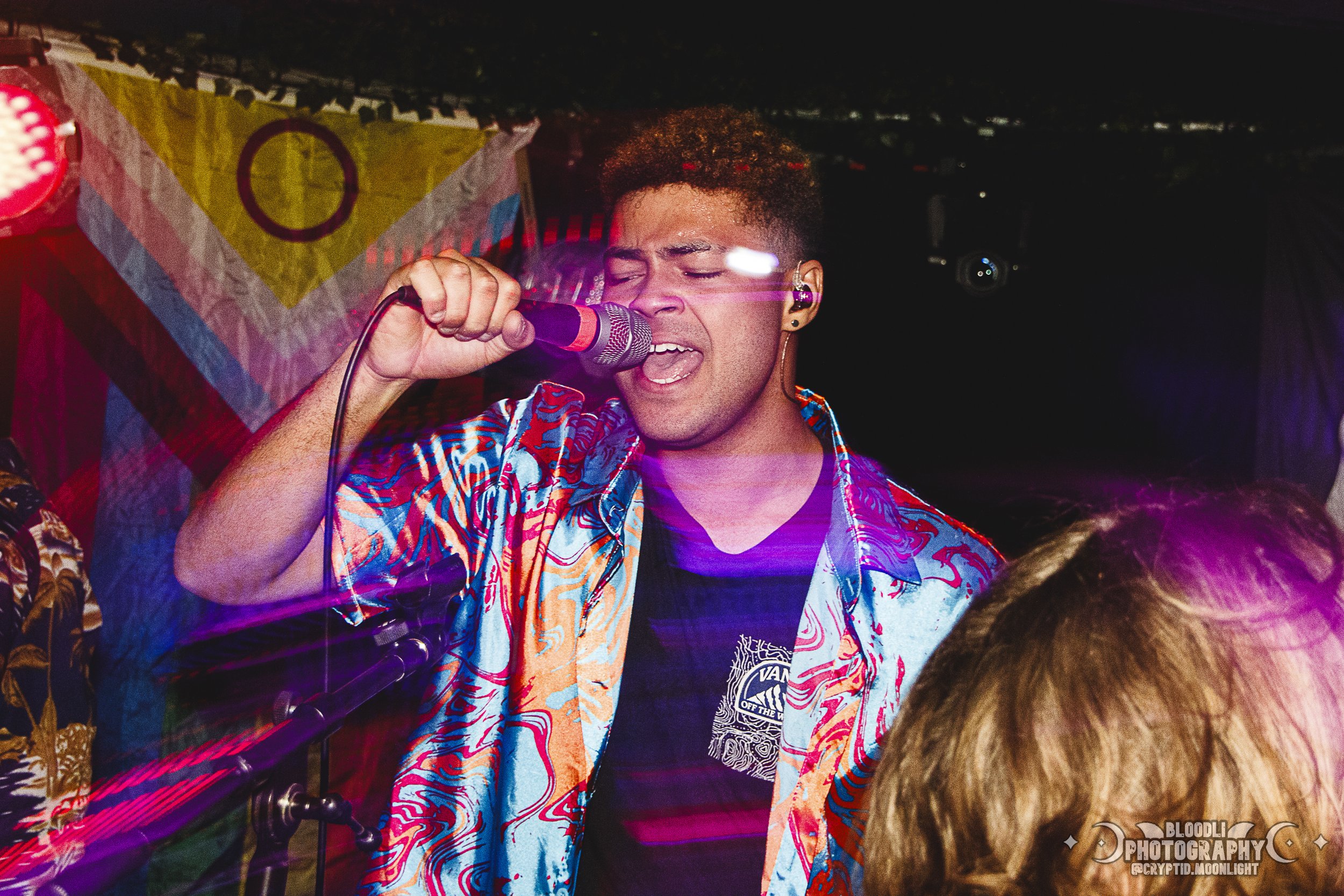 A man passionately singing into a microphone at a lively event, wearing a colorful patterned shirt and a black t-shirt. The background features vibrant lighting and hanging decorative flags.