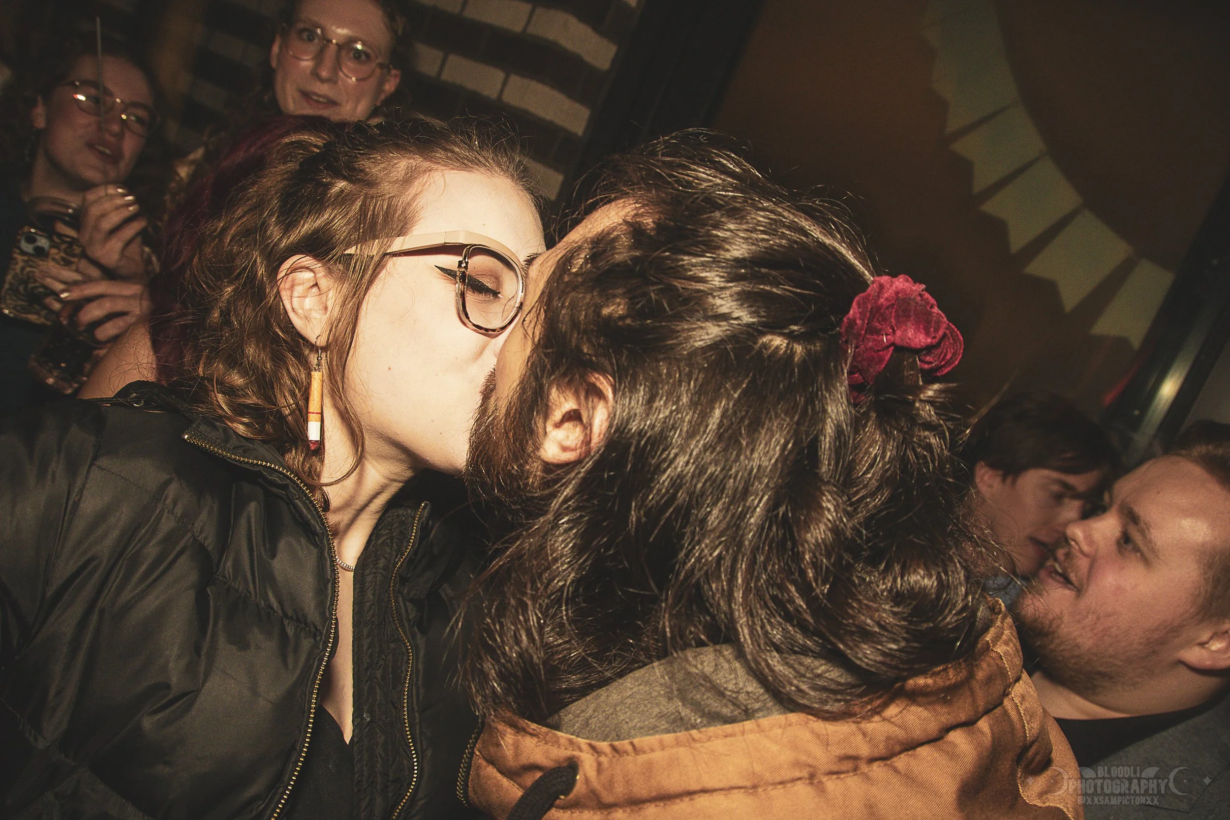 A couple is sharing a kiss on a crowded social gathering. The woman wears glasses and has wavy hair, while the man has dark hair and a beard. People are looking at them and enjoying the moment in the background.
