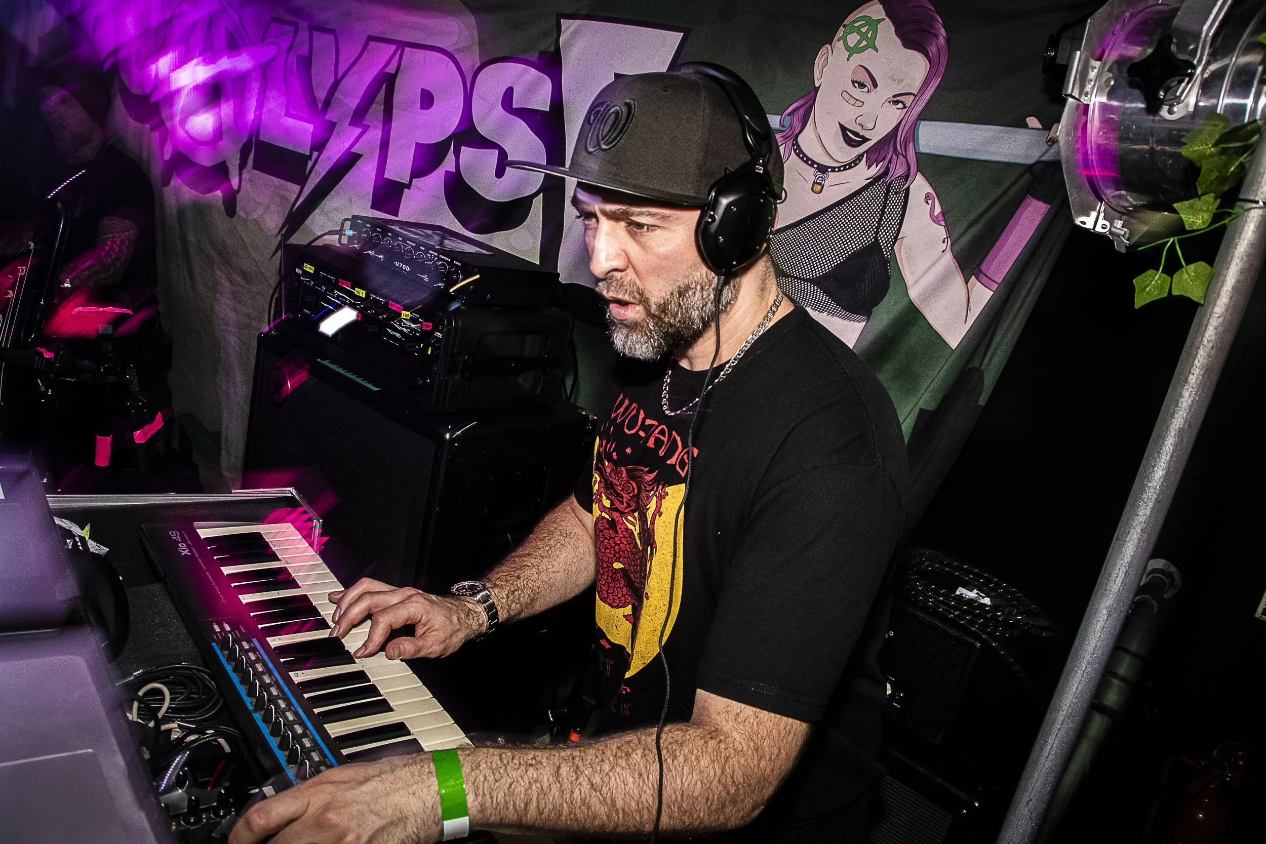 A DJ with a gray beard and black t-shirt playing electronic music at a club, with a keyboard, audio equipment, and a person holding a camera in the background, illuminated by pink and purple lights.
