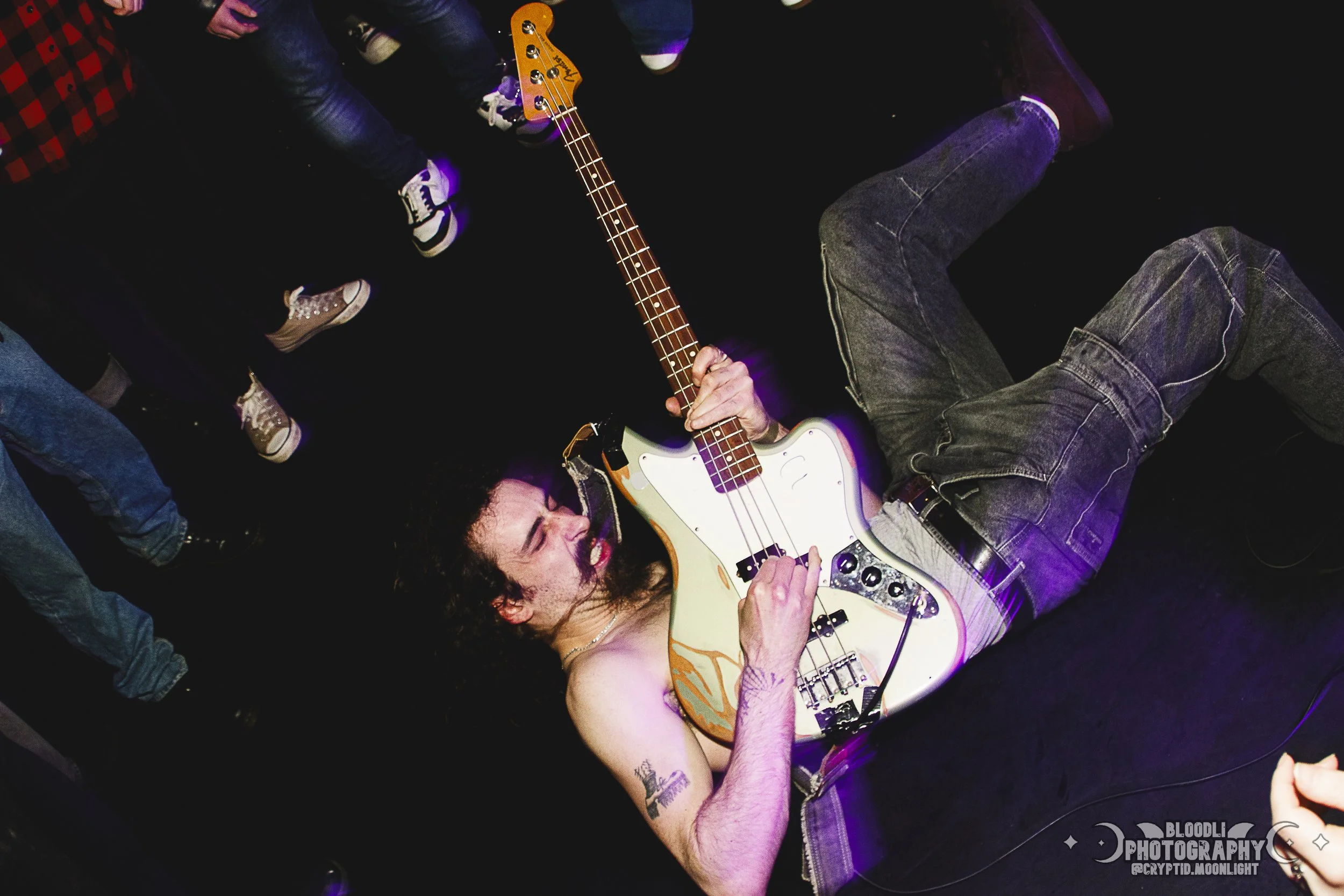 A shirtless man with long hair playing an electric bass guitar on stage during a live music performance. The man is lying on his side, holding the guitar, with people in sneakers and jeans surrounding him or watching from the side.