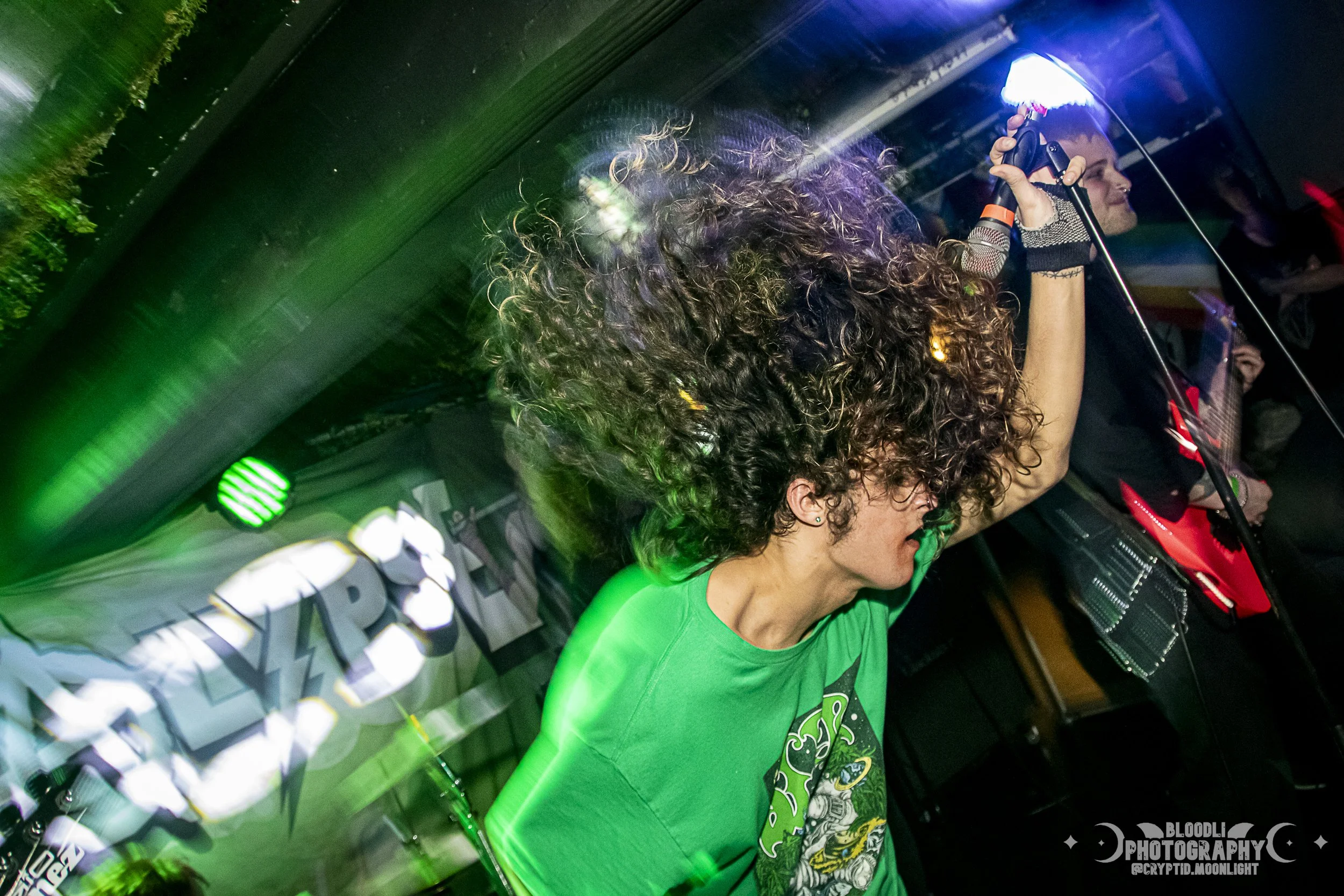 A woman with curly hair wearing a green shirt is dancing with her arm raised, in a nightclub or concert setting with colorful lights.