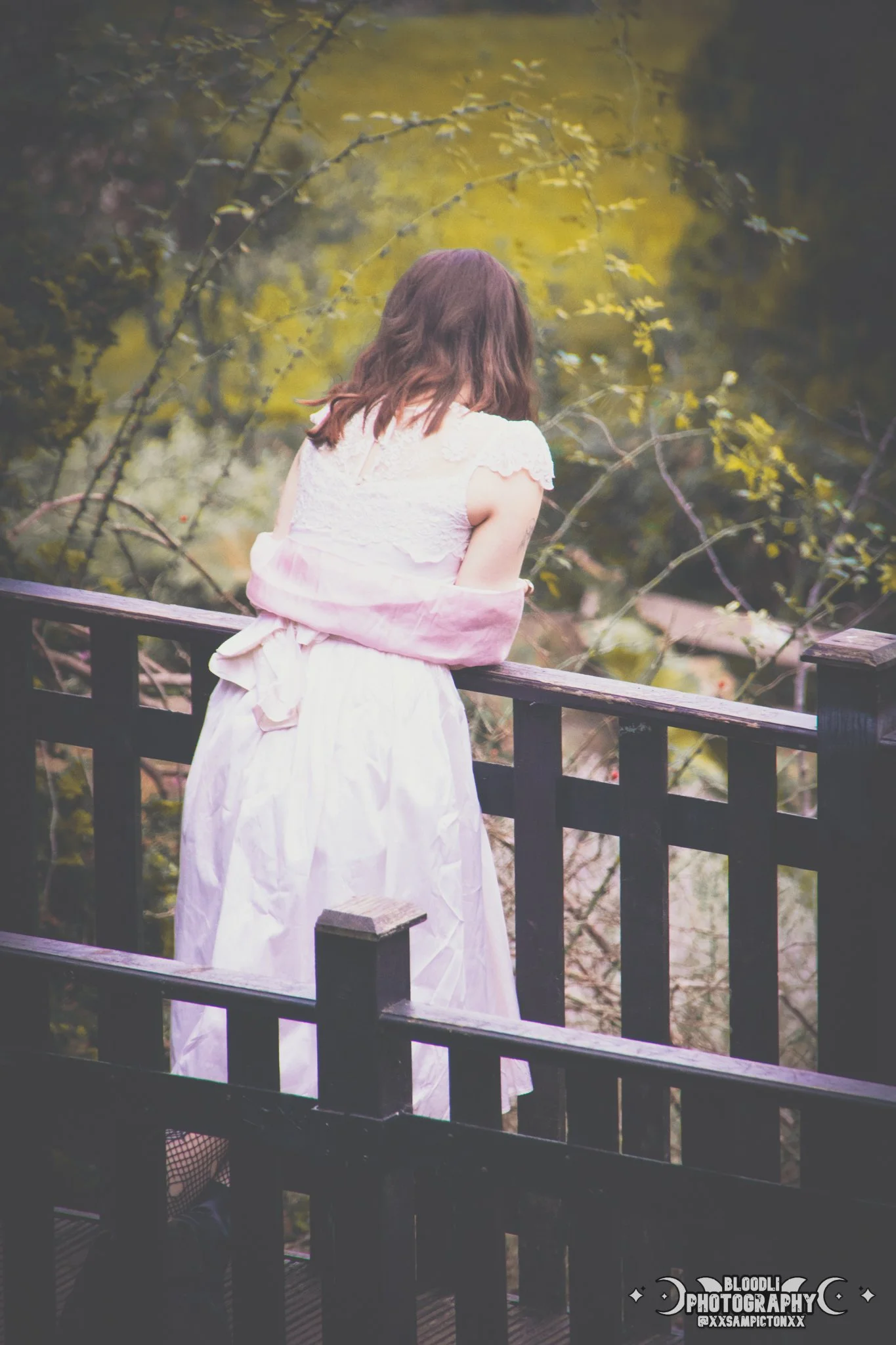 A woman with brown hair stands on a wooden bridge, looking out into a wooded area with autumn leaves.
