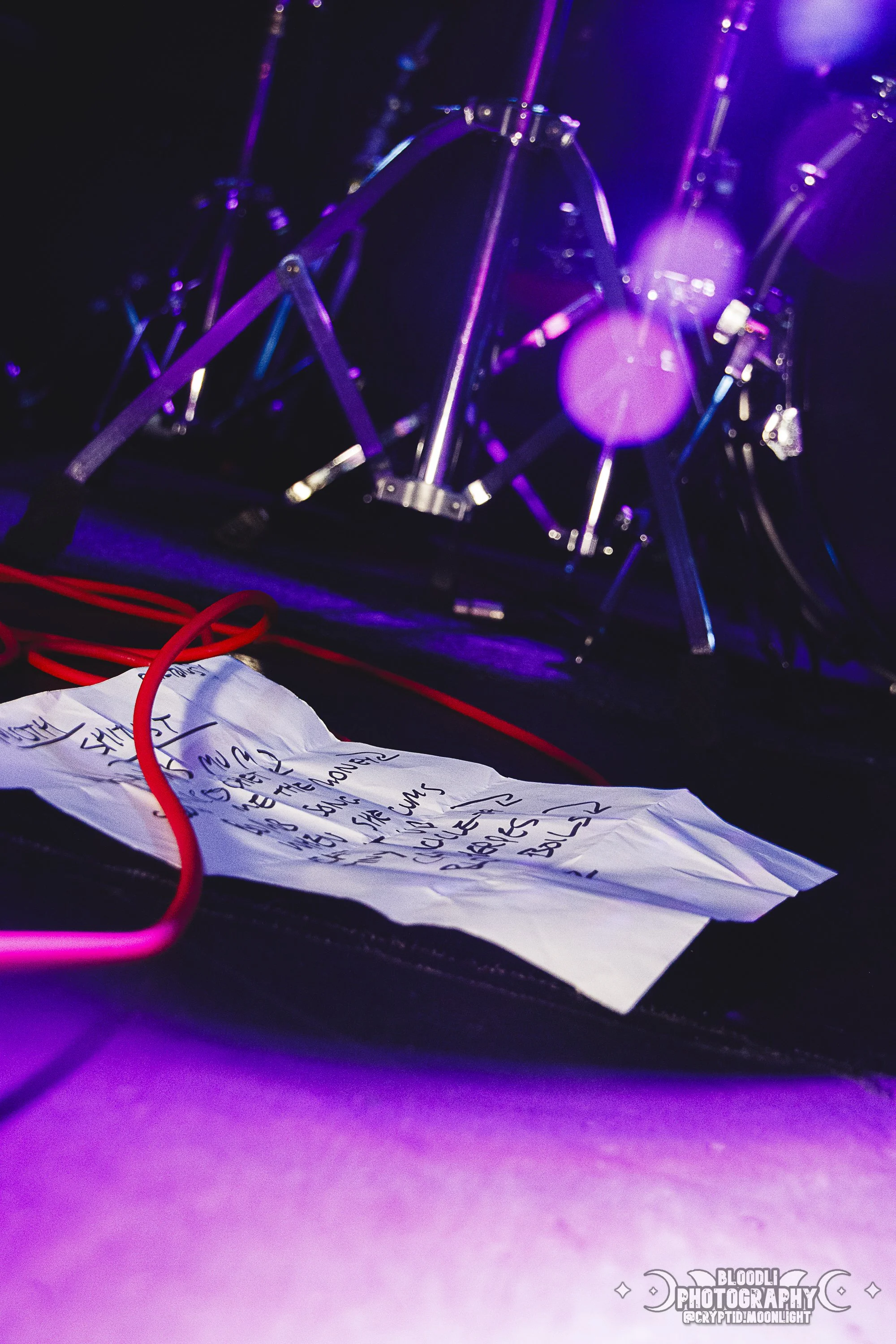 A crumpled handwritten setlist on a stage floor with stage equipment and a music stand in the background, illuminated by purple stage lights.