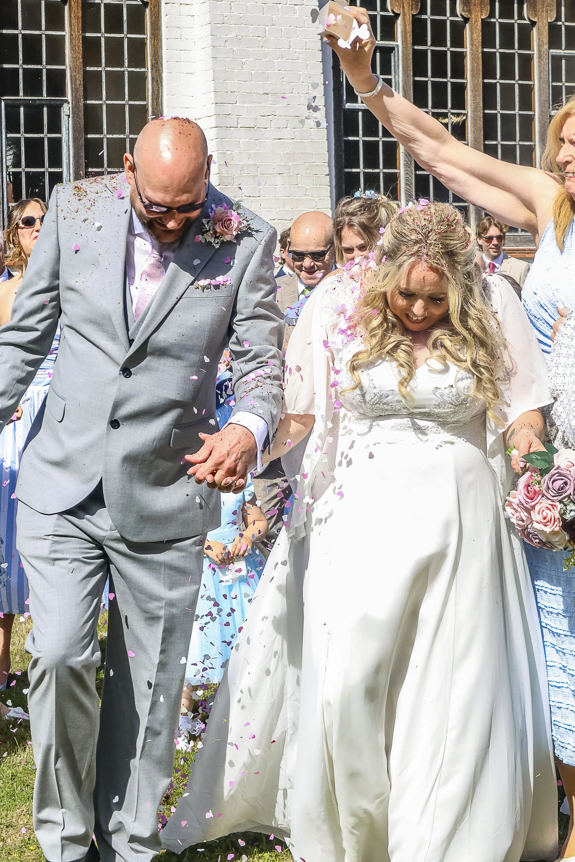 A wedding celebration outside with a man in a gray suit and a woman in a white wedding dress surrounded by guests, confetti, and flowers.