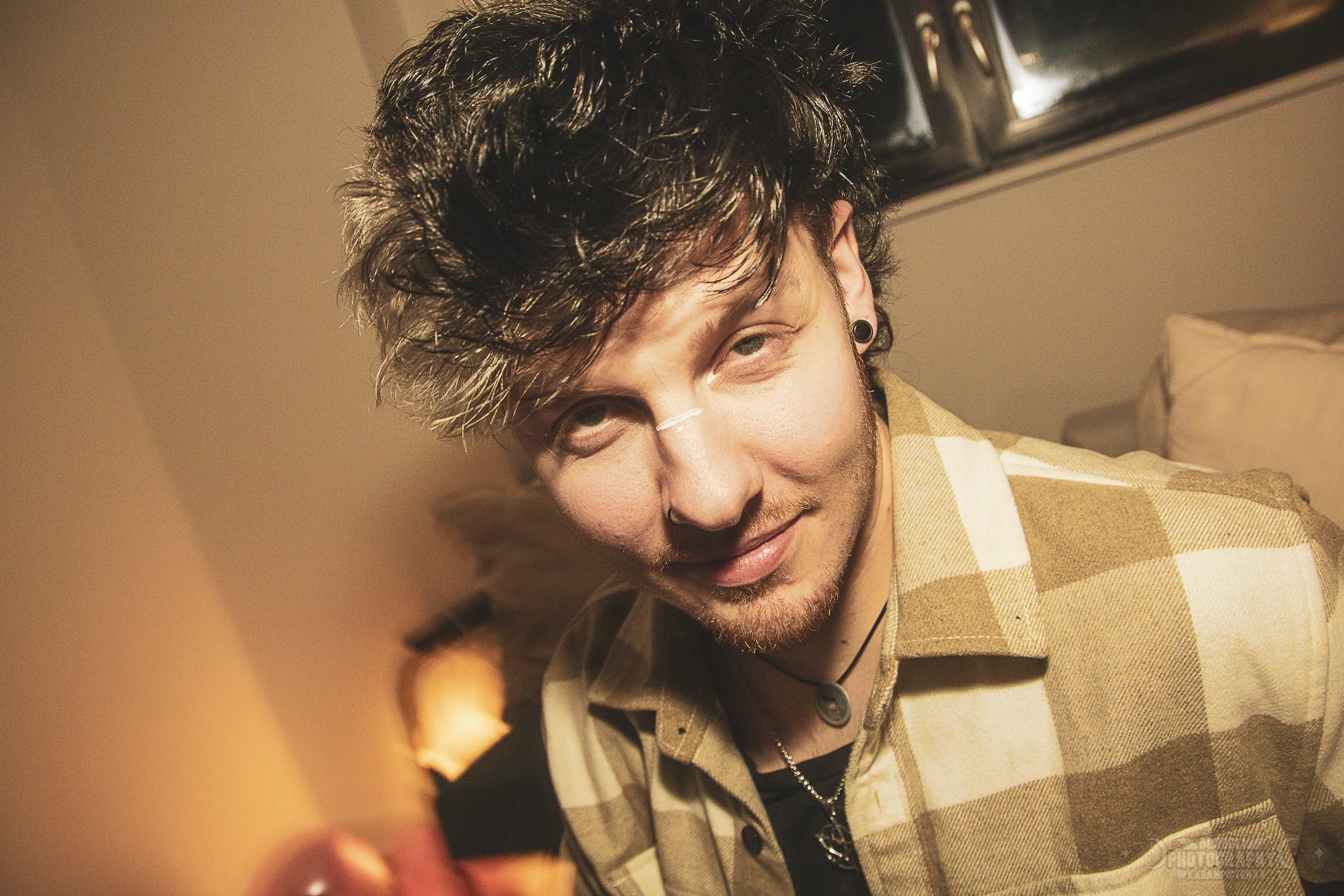 A young man with styled dark hair, ear gauges, and a necklace, wearing a beige plaid shirt, looking at the camera with a slight smile in a warmly lit room.