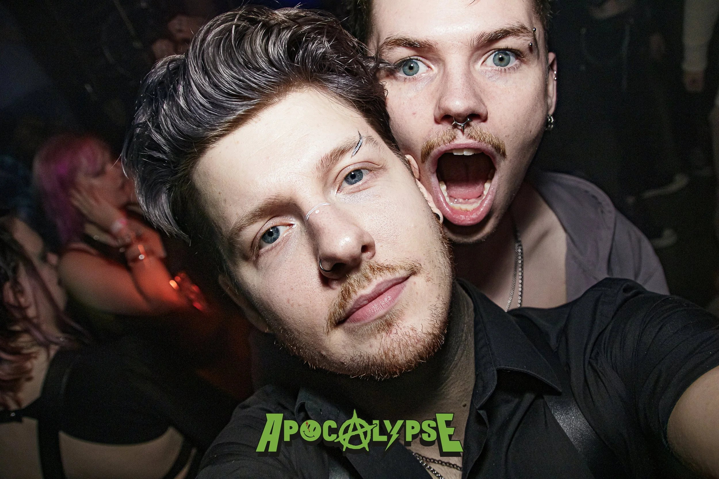 Two young men posing for a selfie at a party, one with dark hair and slicked back, and the other with light hair and multiple facial piercings, with a crowd in the background and the word 'APOCALYPSE' in green text with skull and anarchy symbols at t