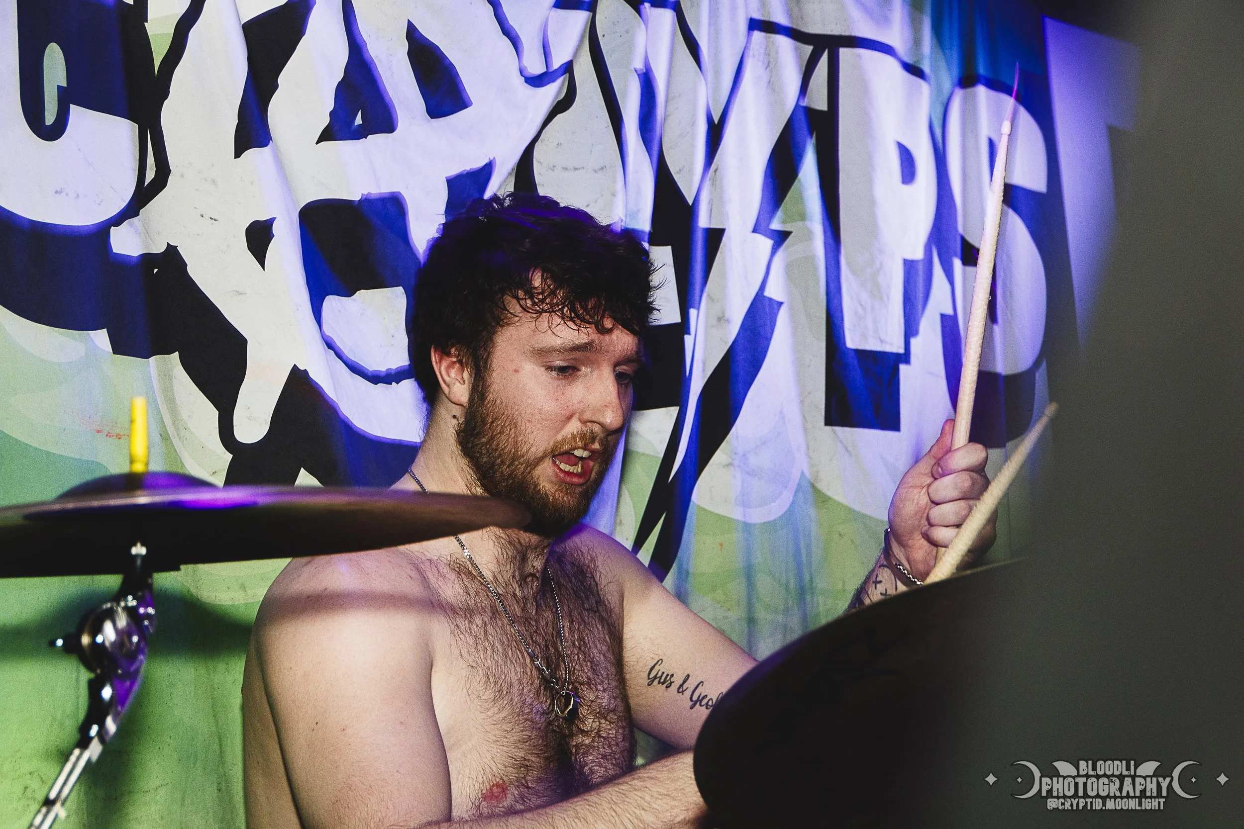 A shirtless young man playing drums at a live music event, with a colorful banner behind him.