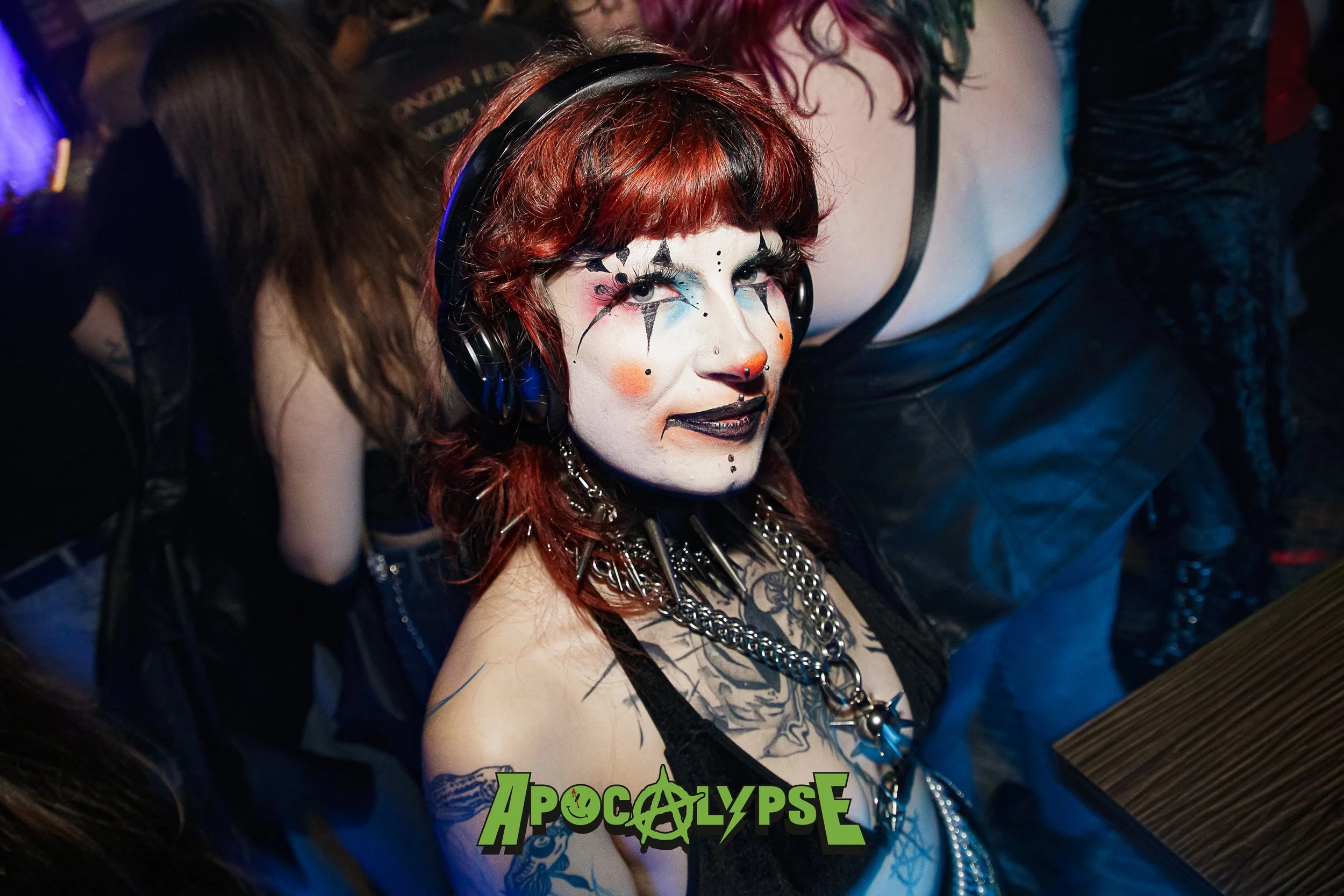 Person with red hair wearing black lingerie, heavy makeup, and punk accessories in a crowded nightclub.