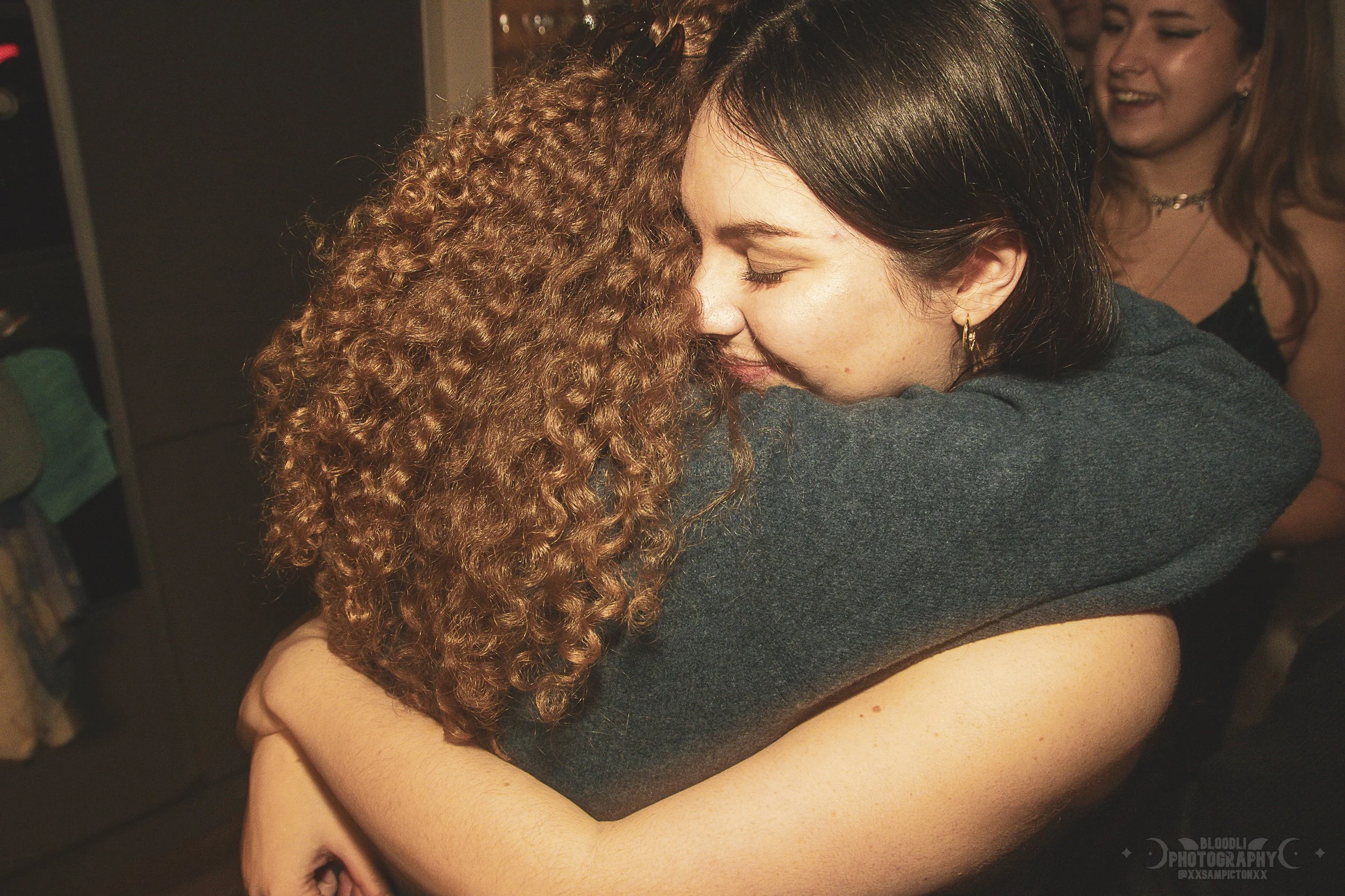 Two women hugging each other tightly with closed eyes, one woman has curly red hair and the other woman has straight black hair. People are in the background smiling and chatting.