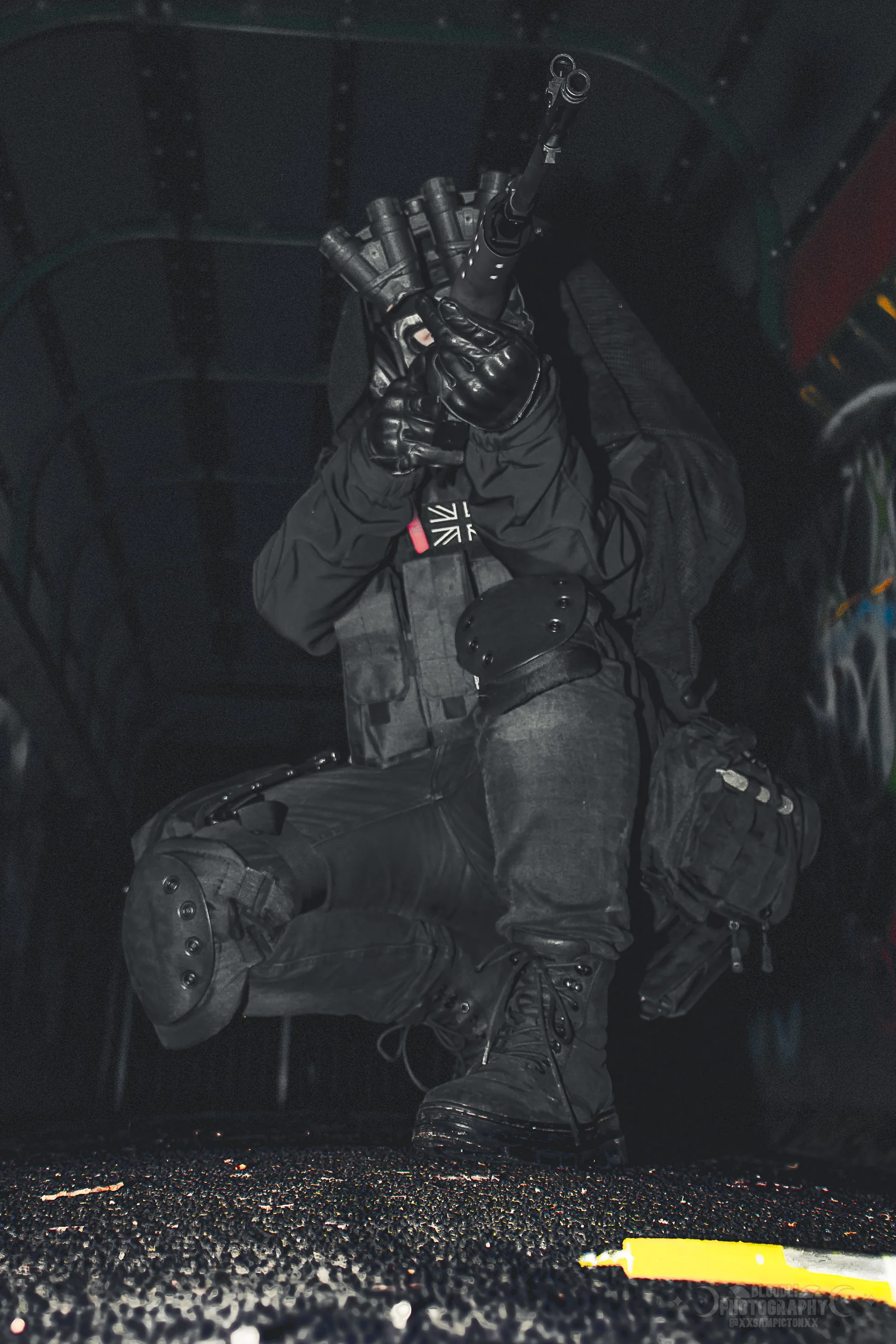 A person dressed in black tactical gear, with gloves and boots, crouching and aiming a rifle inside a dark enclosed space.