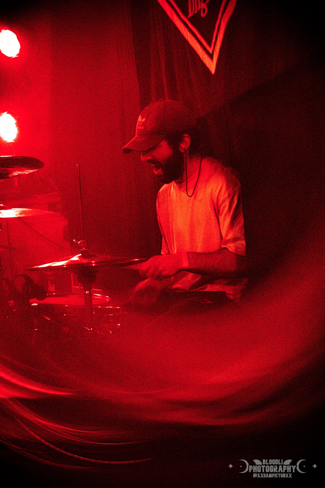 A man wearing a baseball cap and earphones plays drums in a red-lit setting.