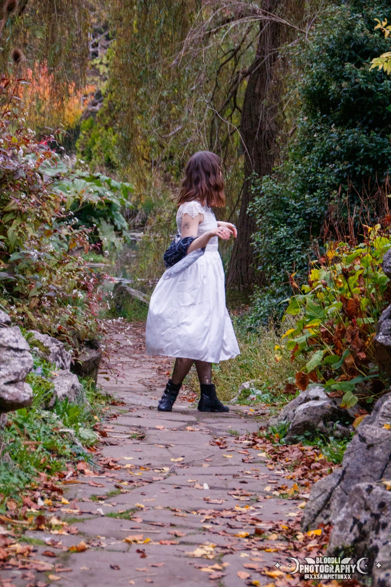A young woman with shoulder-length brown hair, wearing a vintage-style white dress with lace details and black boots, walking along a winding stone pathway surrounded by colorful autumn foliage and trees.