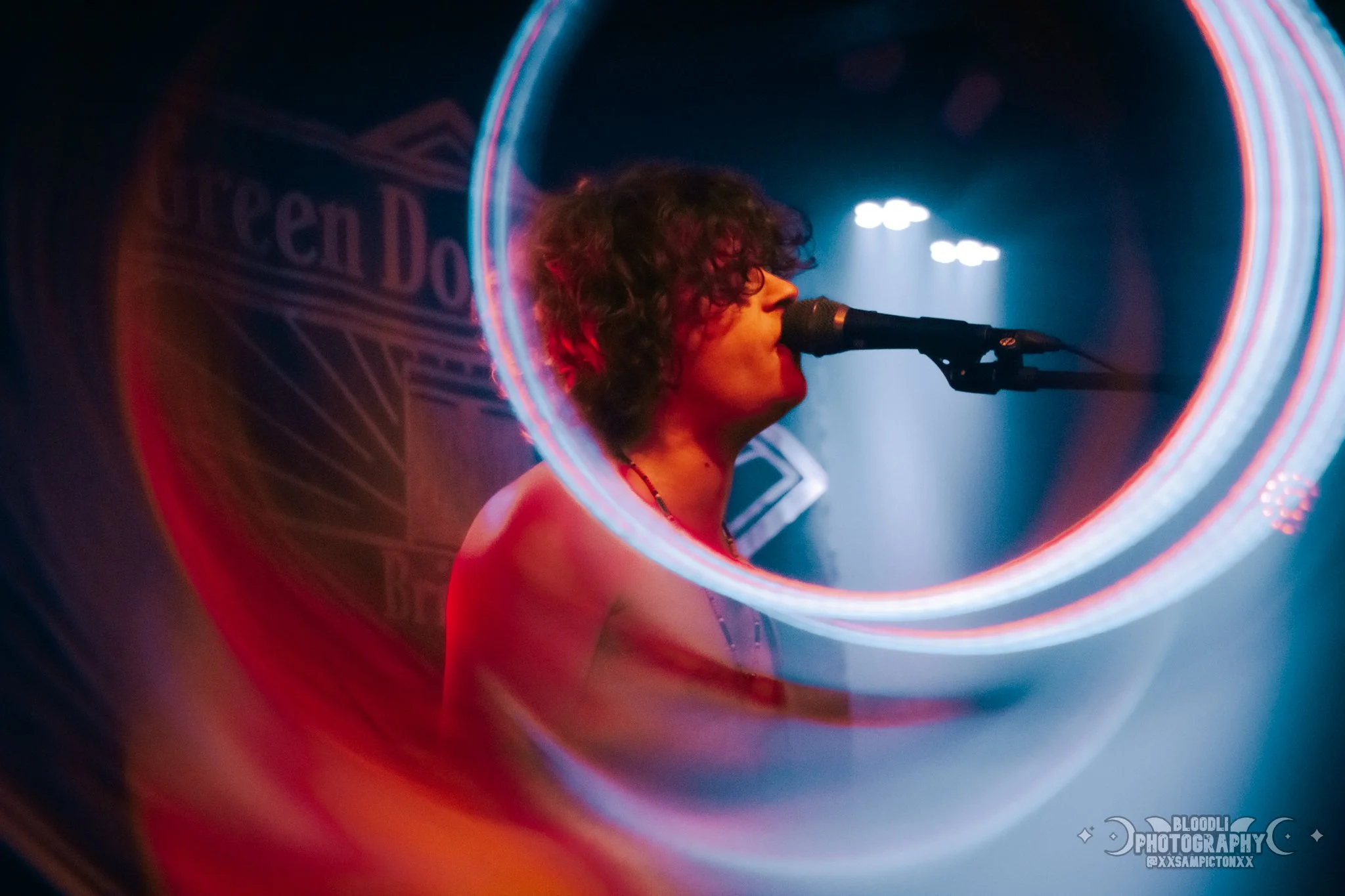 A person with curly hair singing into a microphone on stage with neon lights and a sign that reads 'Green Dot' in the background.