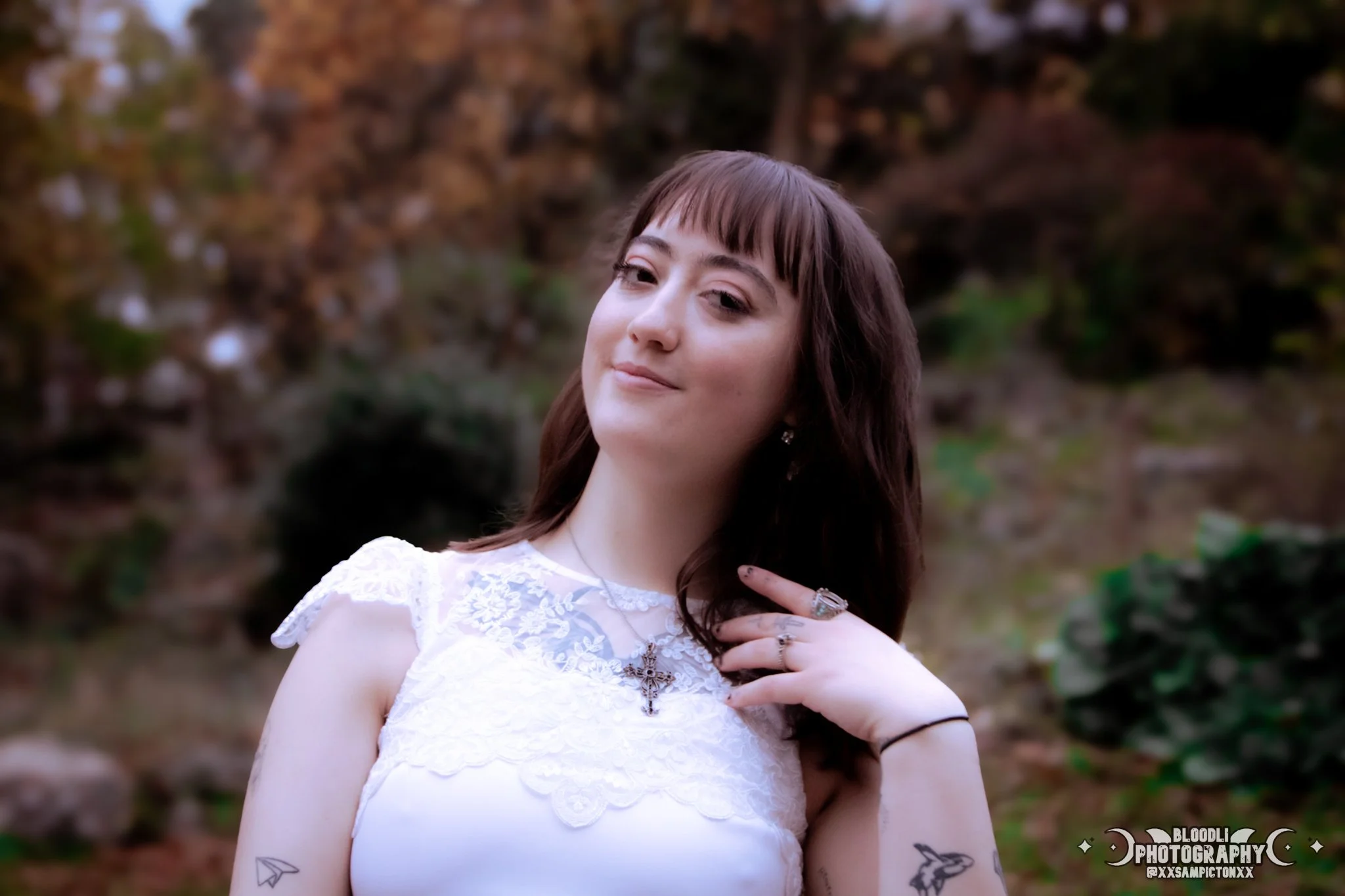 A young woman with brown hair and bangs, wearing a white lace dress, standing outdoors with a blurred natural background, smiling slightly and touching her neck with her right hand, which has rings and a black hair tie.