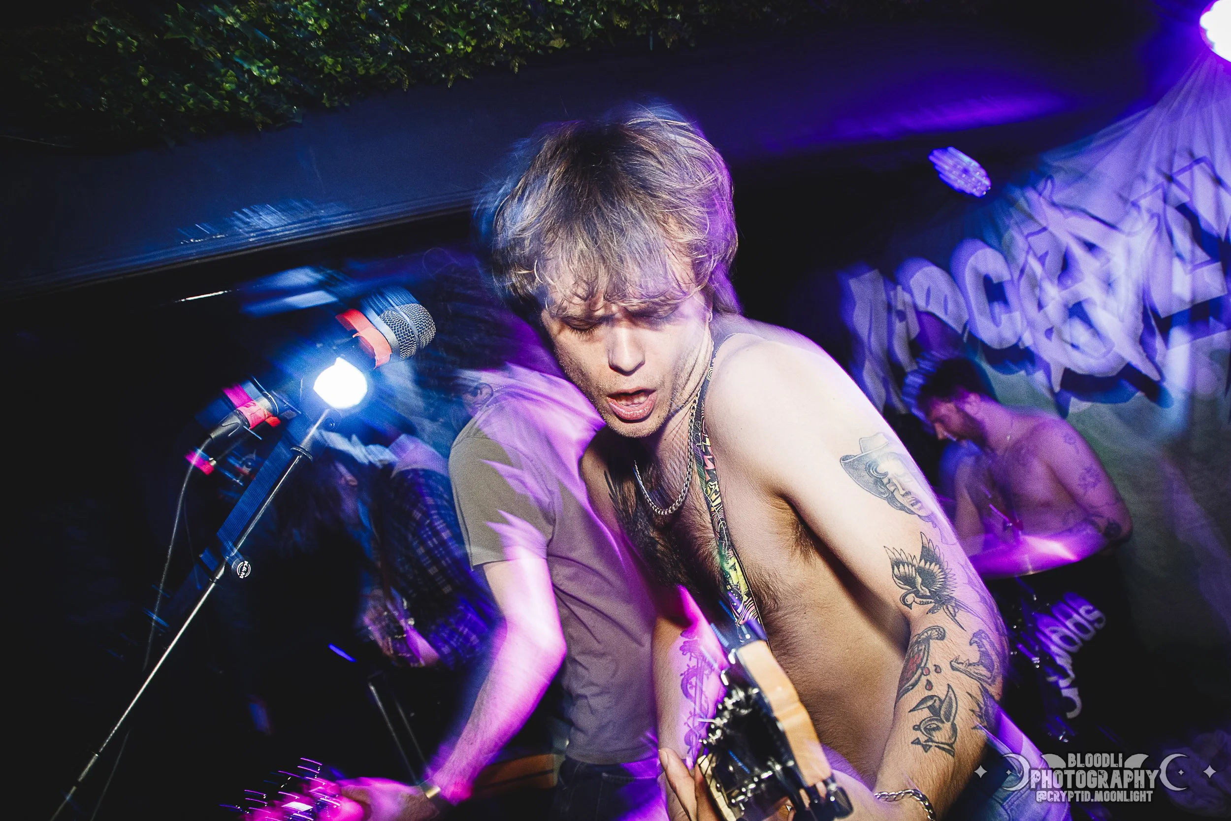 A shirtless male musician with tattoos playing guitar on stage at a concert with colorful lighting and a crowd in the background.