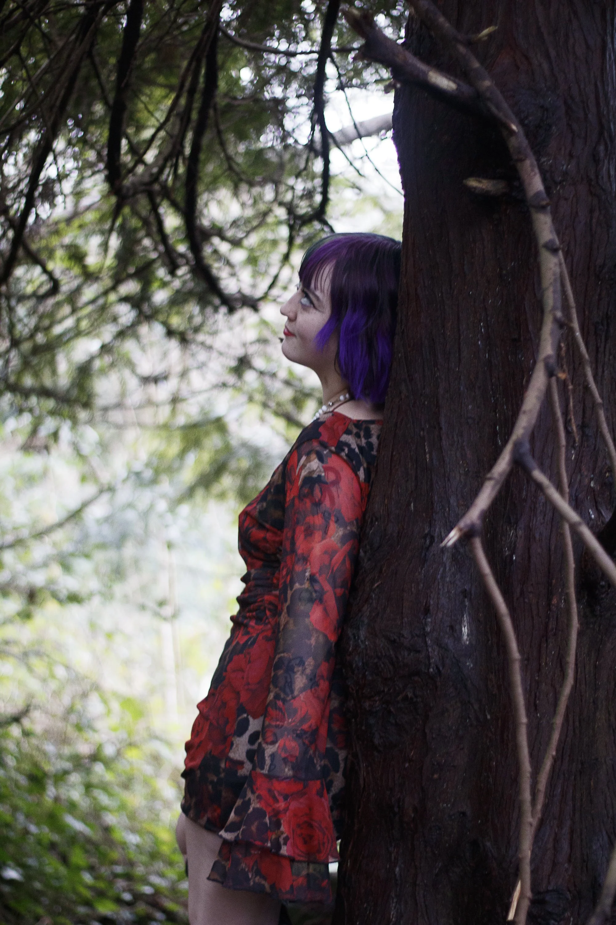 A woman with purple hair wearing a red and black floral dress, leaning against a large tree in a forested area.