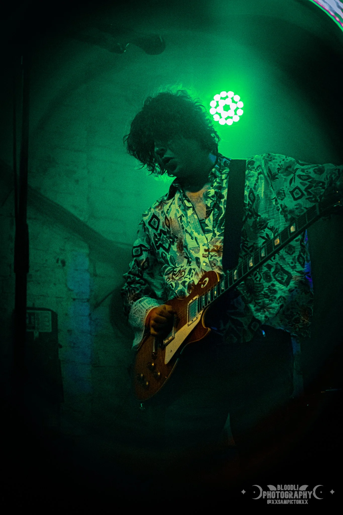 A musician with curly hair playing an electric guitar on a dimly lit stage with green lights.