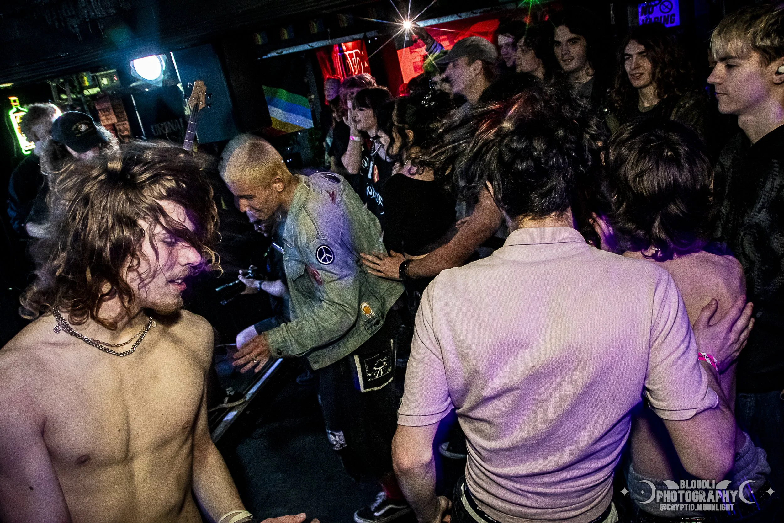 Group of people dancing and socializing in a dark, colorful nightclub with neon lights, some with colorful hair and casual clothing, and one shirtless man with curly hair.