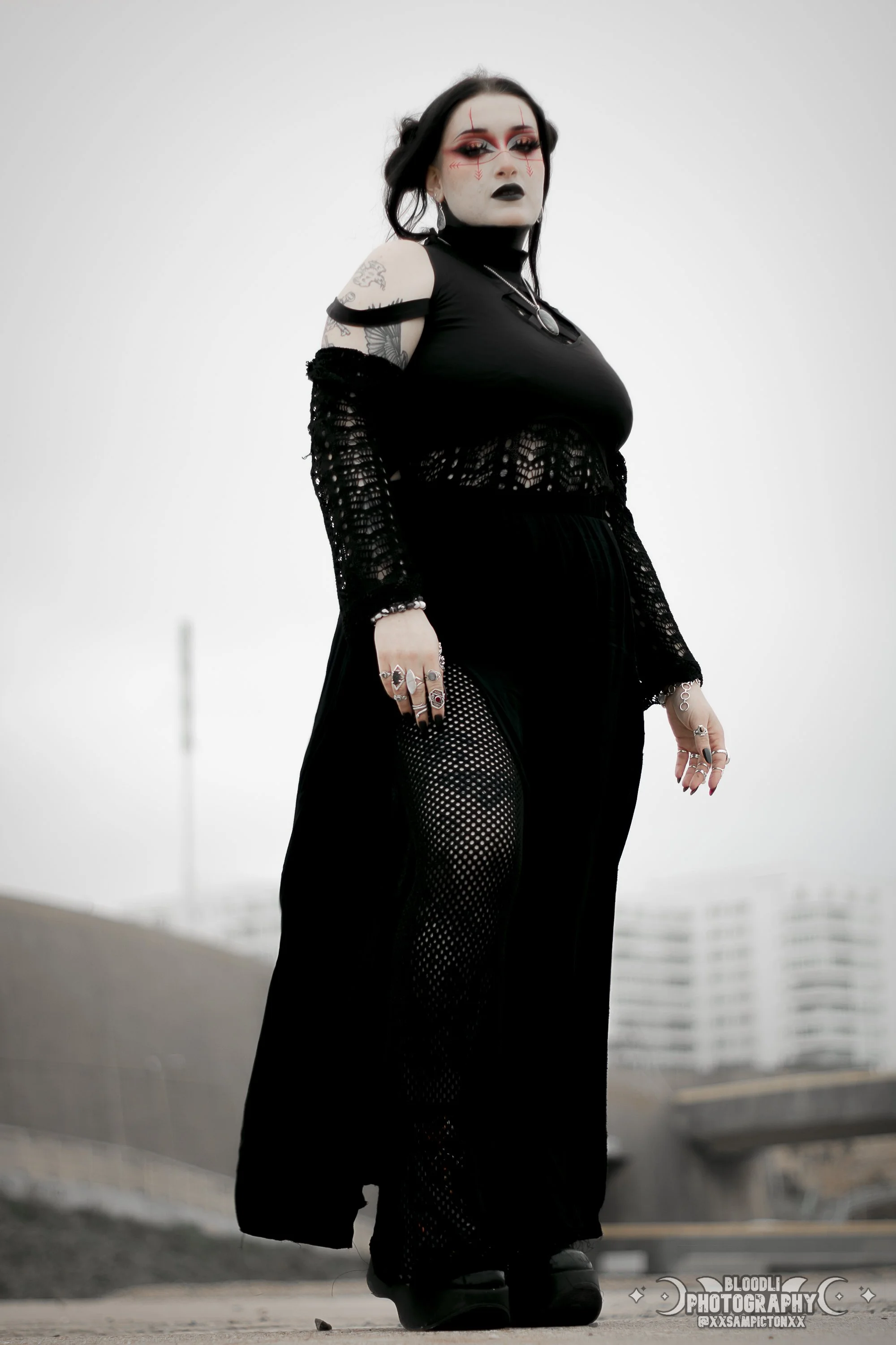 A woman with dark hair, gothic makeup, and tattoos standing outdoors wearing black clothing, fishnet stockings, and various rings and jewelry.