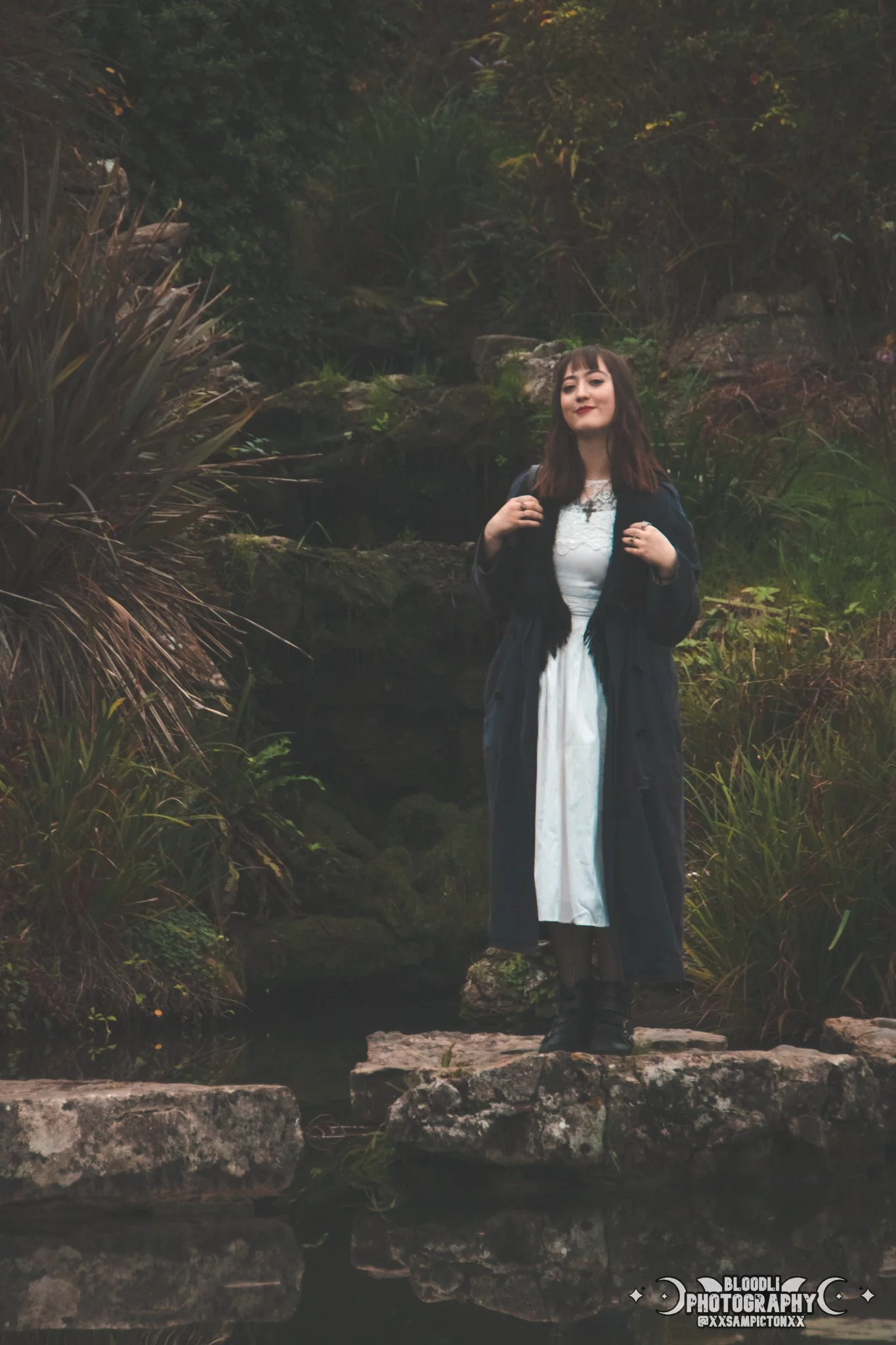 A woman with dark hair, wearing a white dress and black boots, stands on rocks near water in a lush garden with plants and greenery in the background.