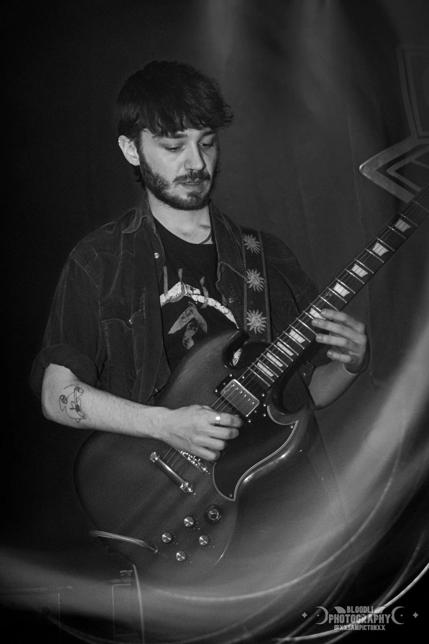 A young man playing an electric guitar on stage, with a focused expression, dark hair, and several tattoos, in a black and white photograph.