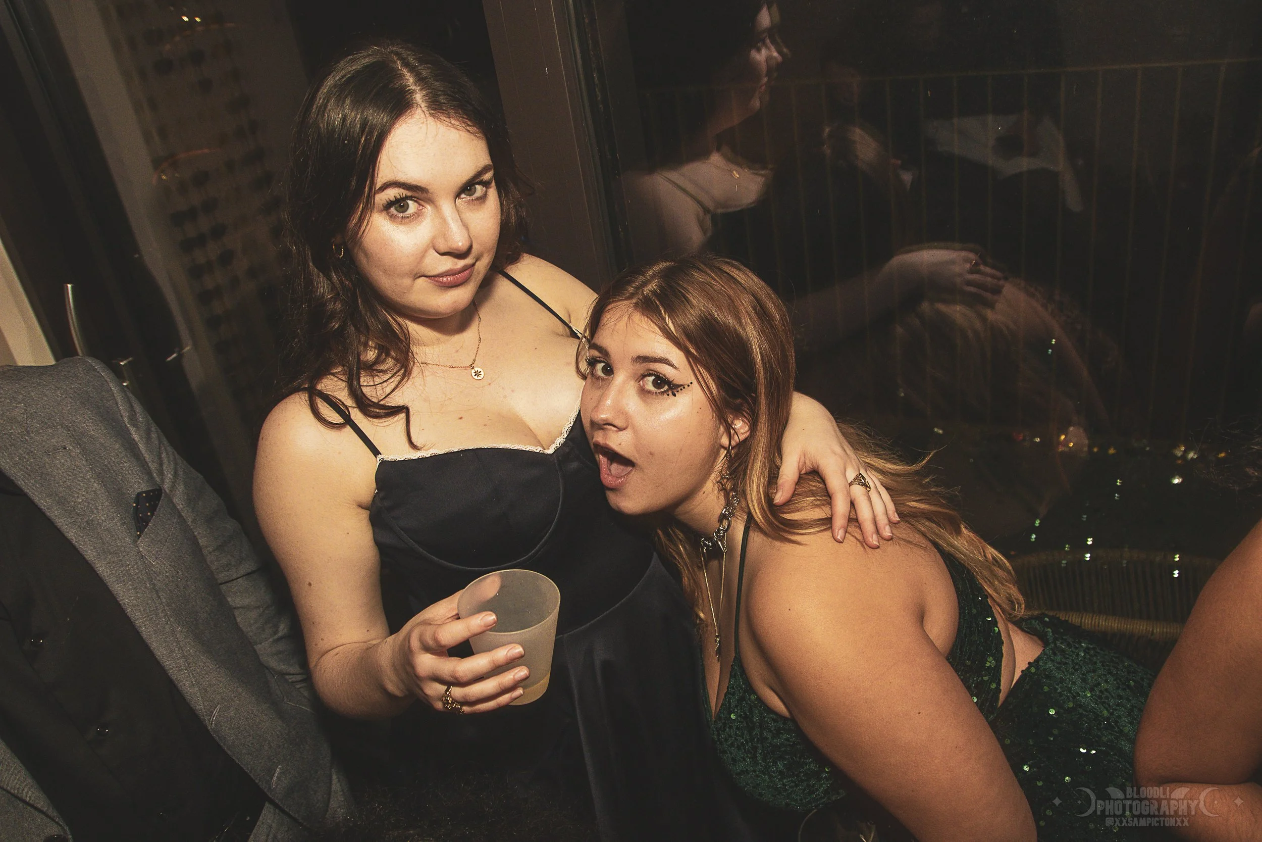 Two women at a party, one holding a drink and the other with her mouth open, posing playfully together.