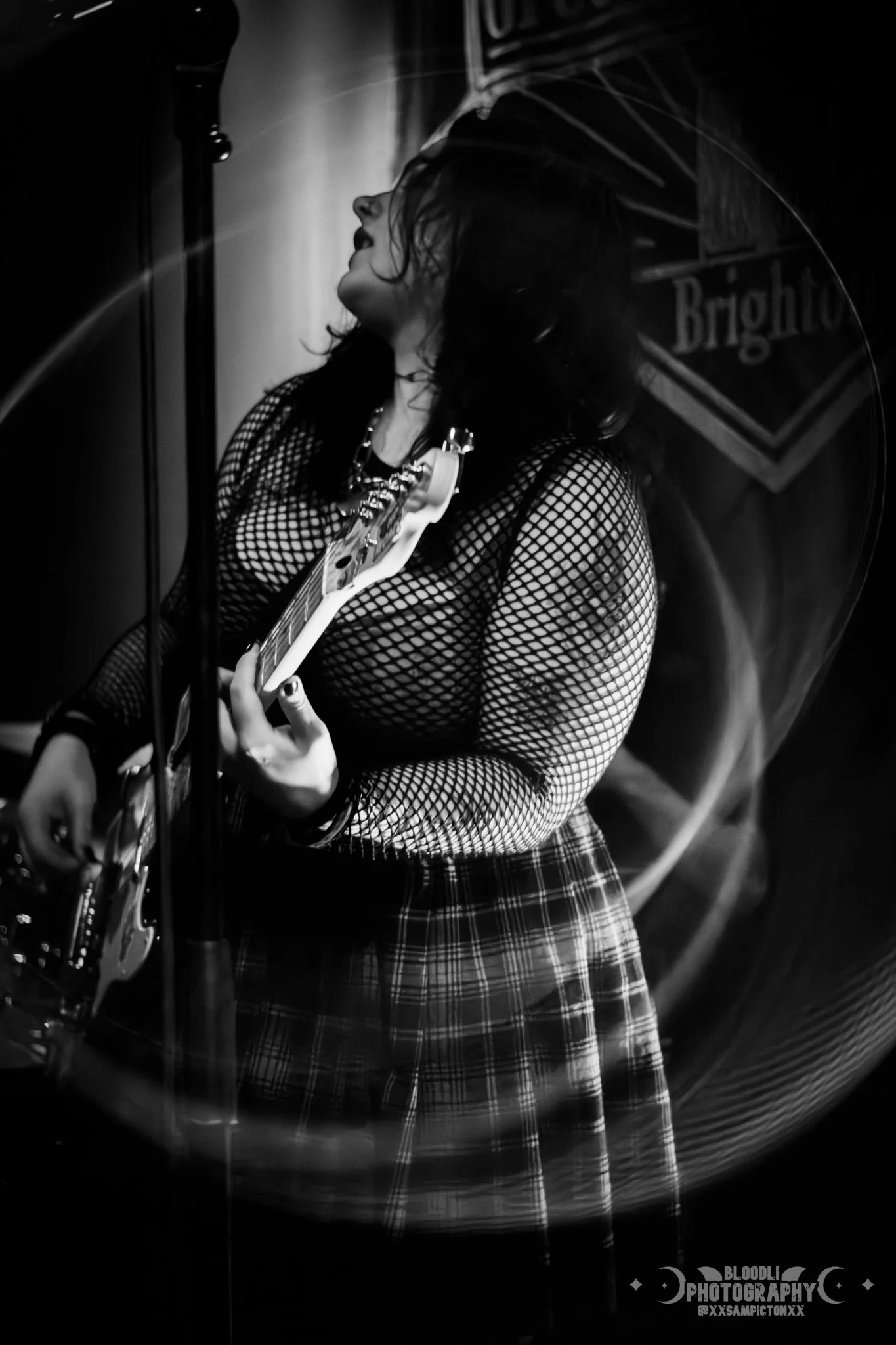 A woman with dark hair playing an electric guitar, dressed in a fishnet top and plaid skirt, captured in black and white, performing on stage.