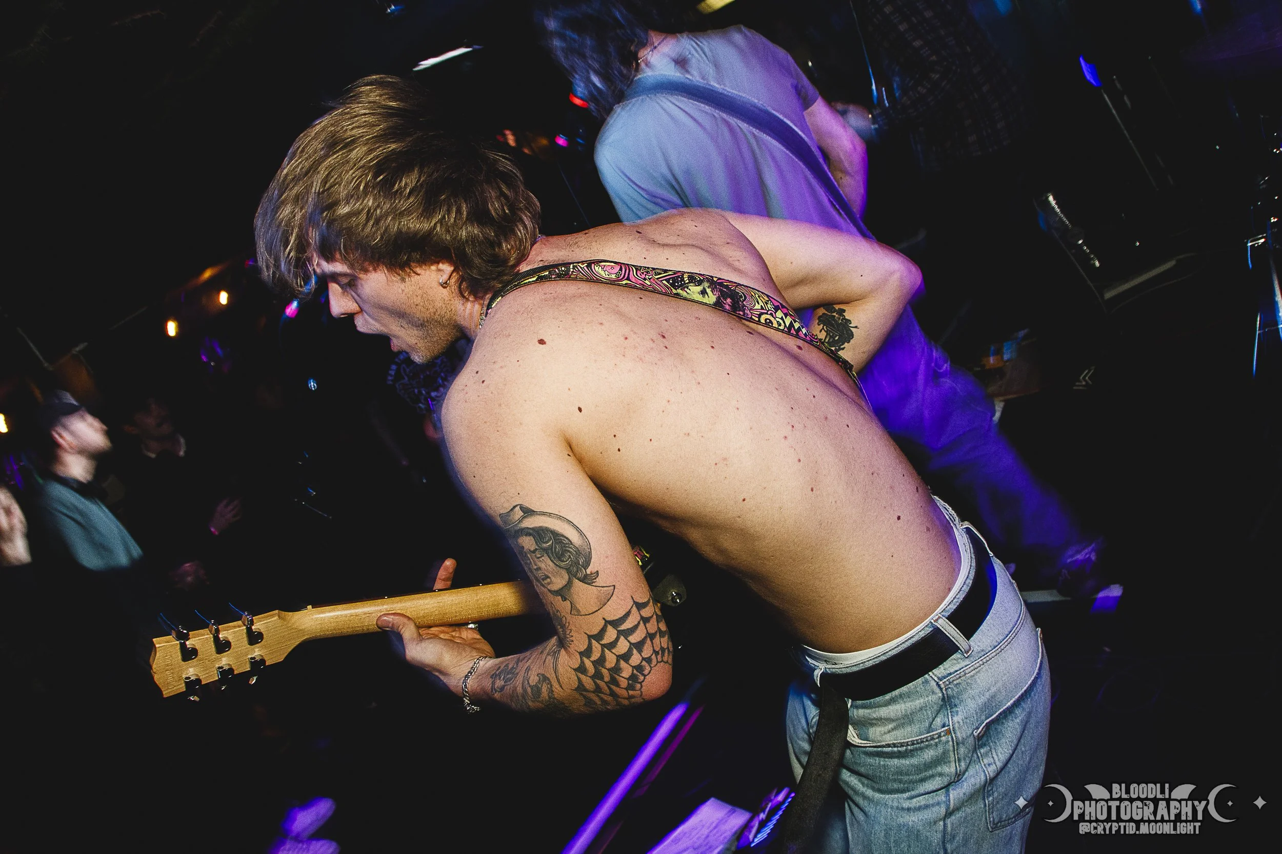 A shirtless male guitarist playing guitar at a concert with people in the background.