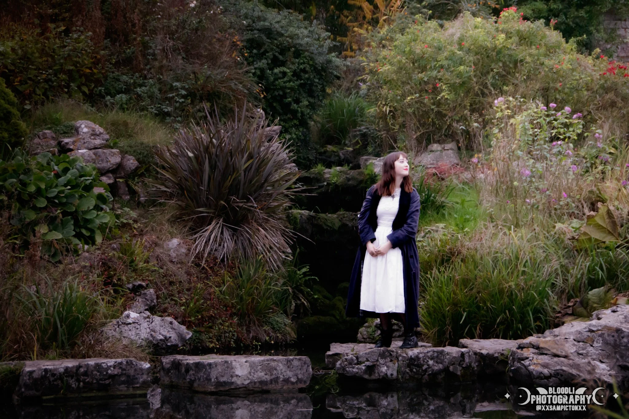 A woman dressed in a white dress and dark coat standing on rocks near a pond, surrounded by lush greenery and flowering bushes.