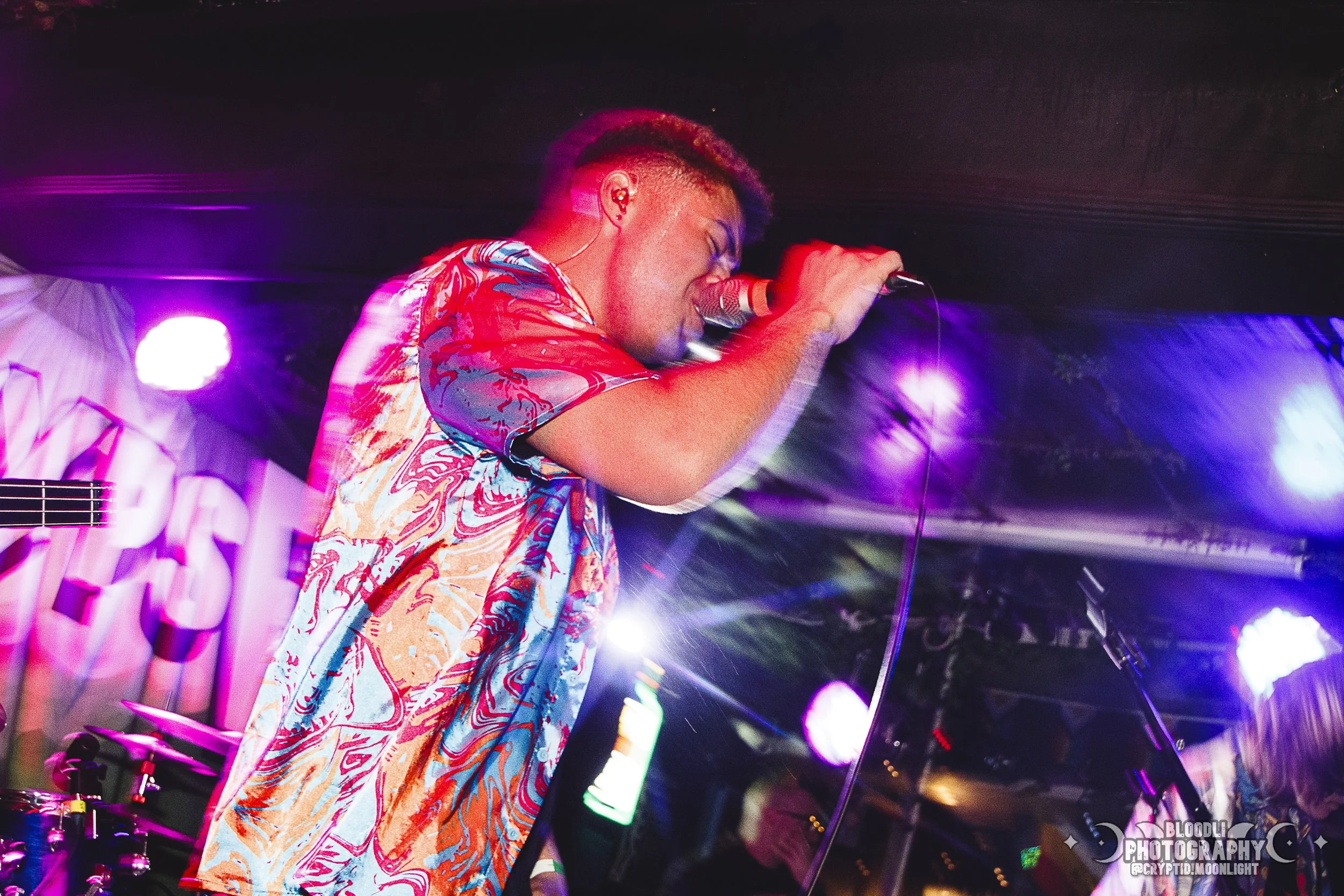 A male singer performing on stage, holding a microphone with his eyes closed, wearing a colorful patterned shirt, under purple and blue stage lights.