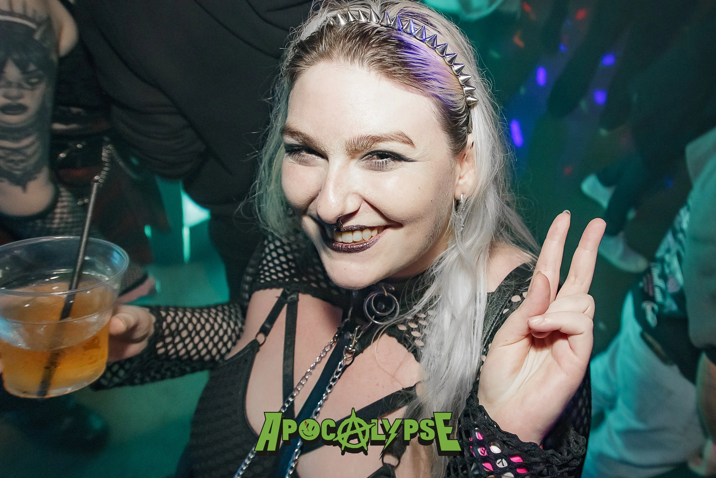 A woman with platinum blonde hair, wearing dark makeup with metallic lipstick, making a peace sign and smiling at a party. She has a spiked headband and is holding a drink. There is a green 'Apocalypse' logo at the bottom.