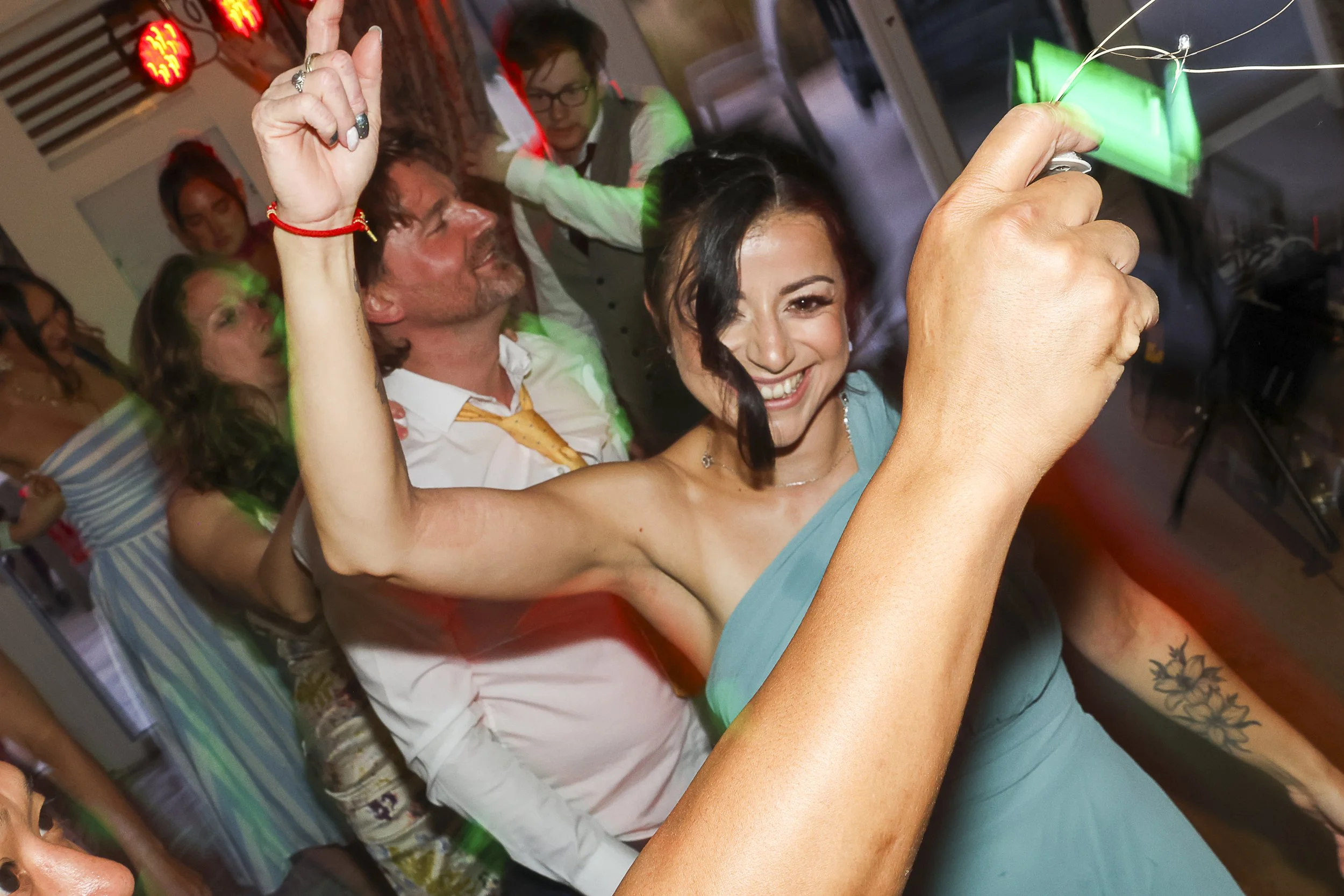 Group of people enjoying a celebration or party, dancing and smiling, with one woman in the foreground holding sparklers. The scene appears lively with colorful lighting.
