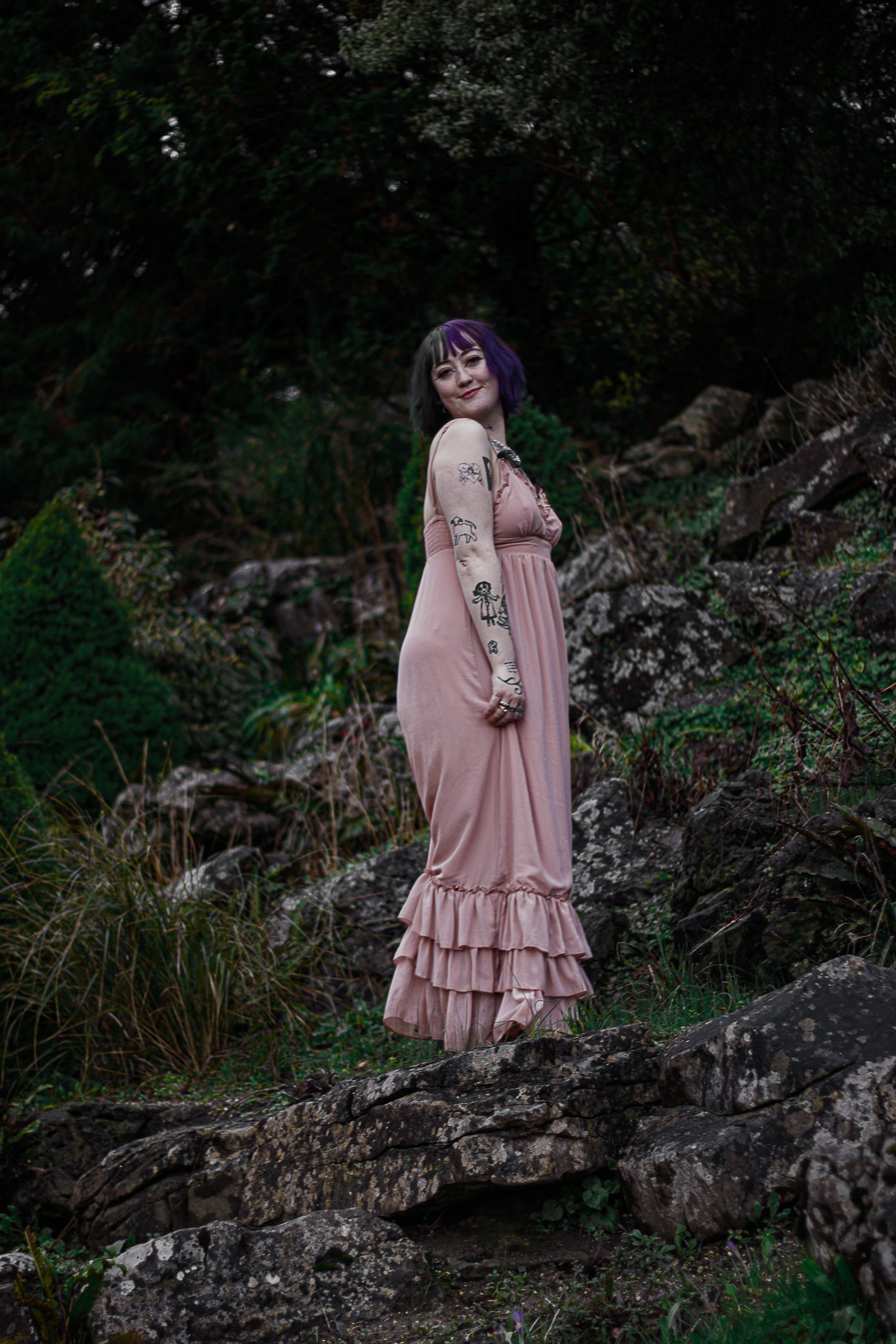 A woman in a pink dress standing on rocks in a natural outdoor setting with greenery and dark trees in the background.