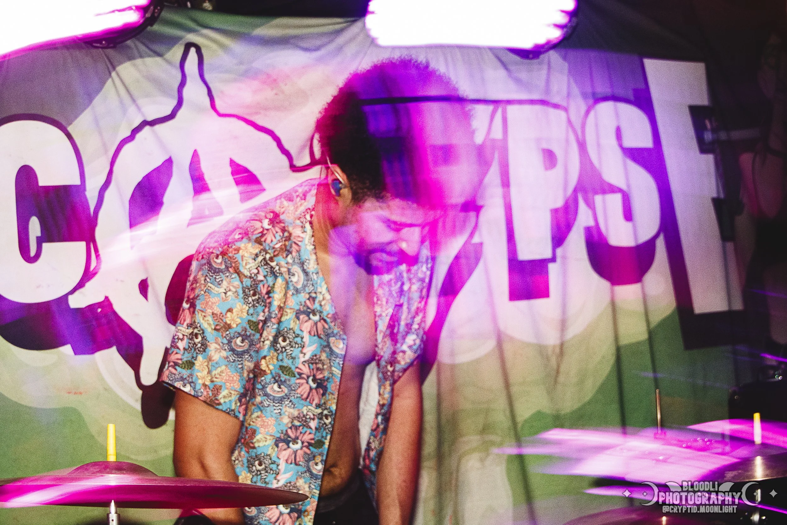 A man performing DJ music at a club with colorful lighting, wearing glasses and a floral shirt, with a large banner in the background displaying a peace symbol and the words 'CURES APPS'.